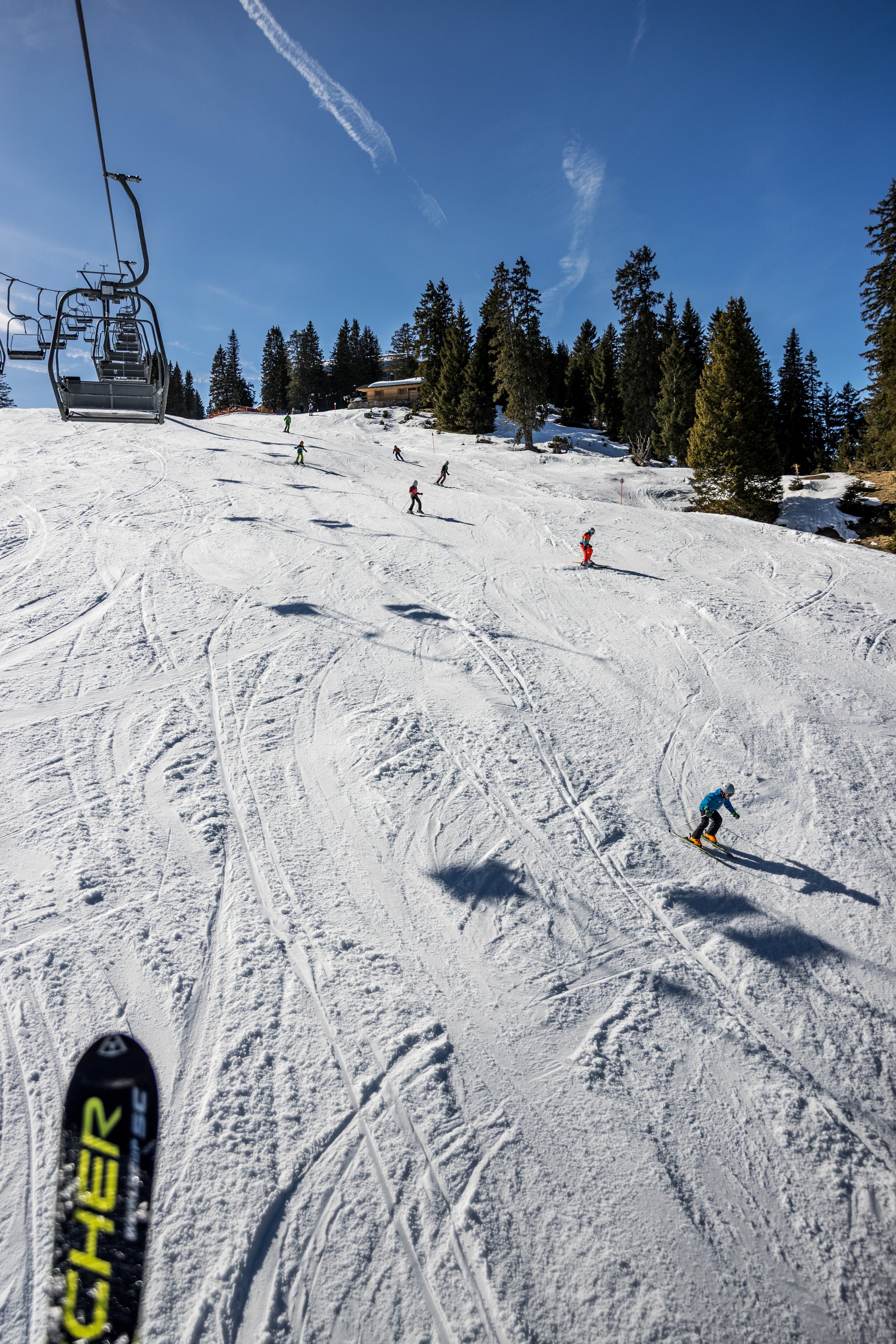 Skiers descending a snowy mountain slope under a clear blue sky, with a ski lift on the left and trees on the right side of the slope.