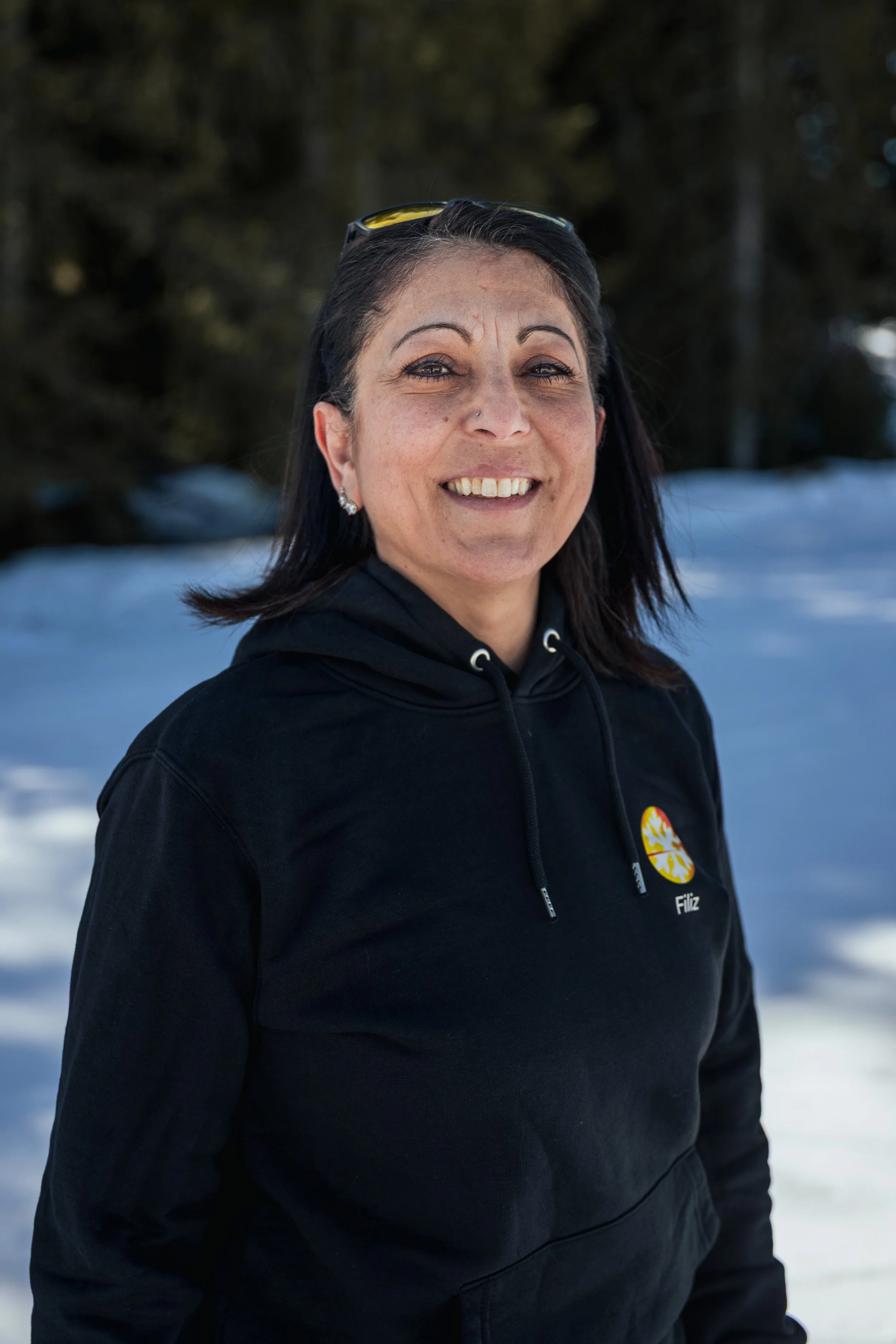 A woman smiling outdoors in a snowy landscape with trees in the background, wearing a black hoodie with a logo on it.