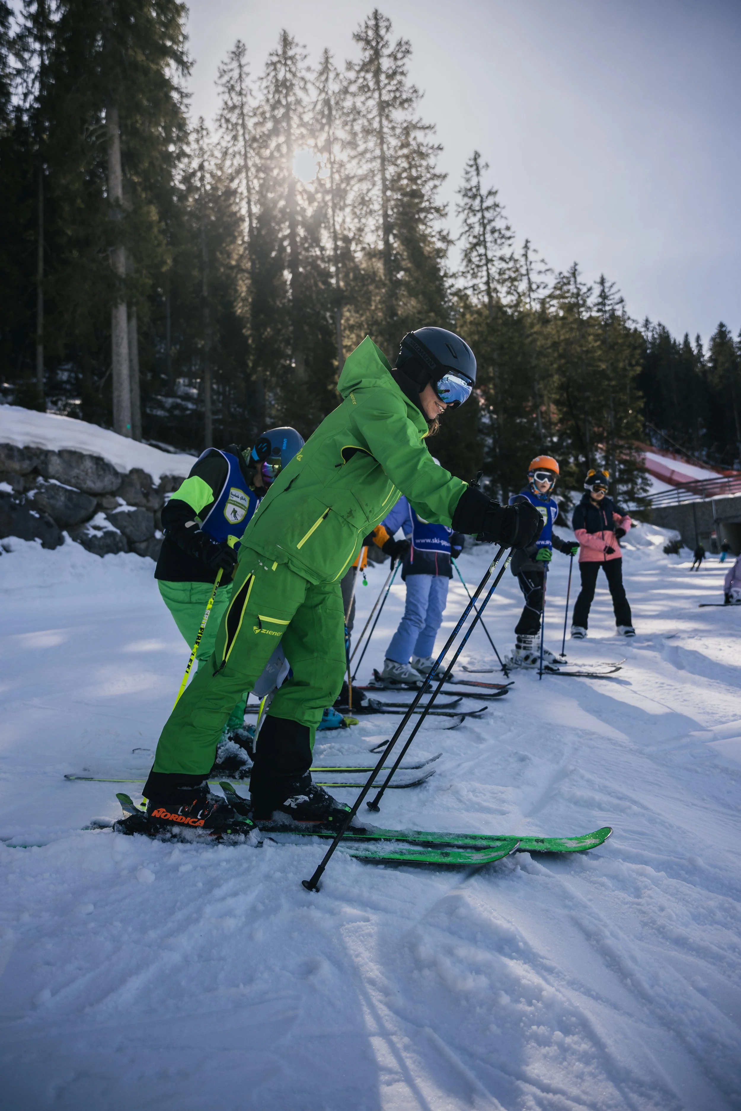 Group of skiers in colorful winter gear, standing on snow with ski poles, preparing for downhill skiing at a mountain resort with trees and a ski lift in the background.