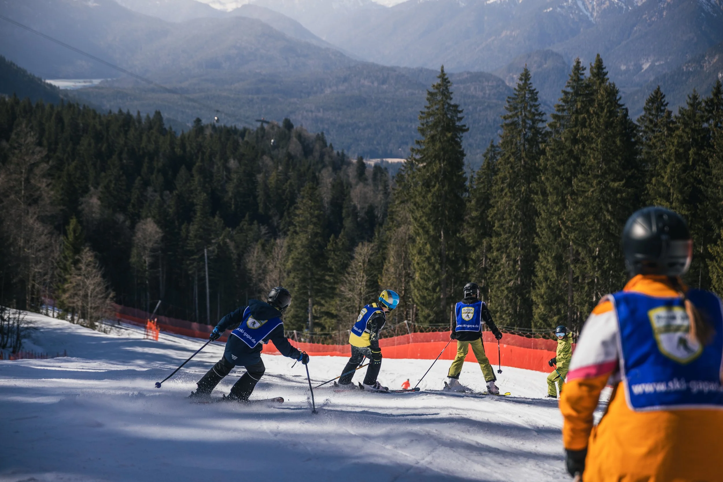 Children skiing on a snow-covered mountain with a backdrop of tall evergreen trees and distant mountains, some wearing helmets and colorful winter gear.
