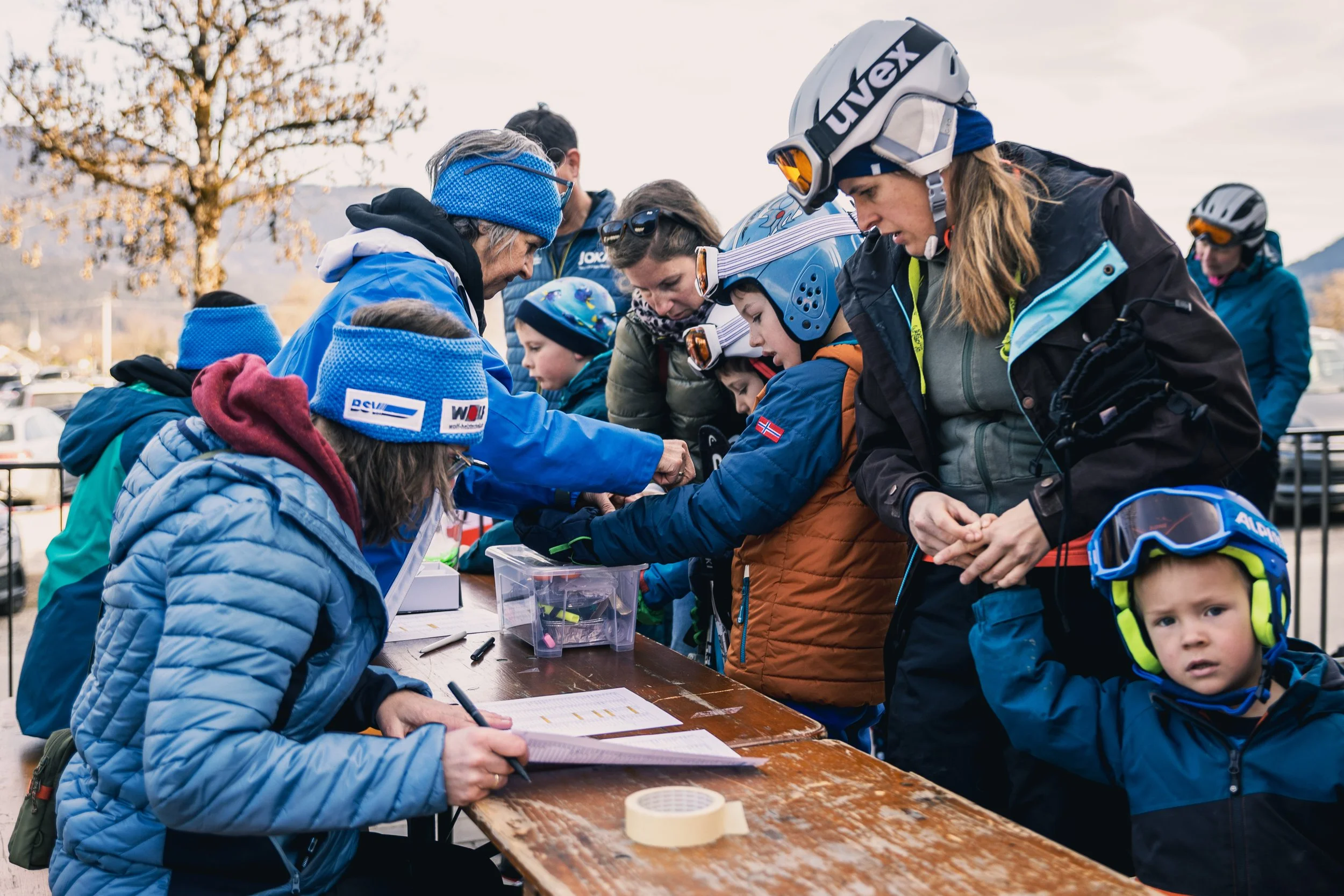 Group of children and adults, all wearing winter and ski gear, gathered around a table outside registering or checking in for a skiing event.