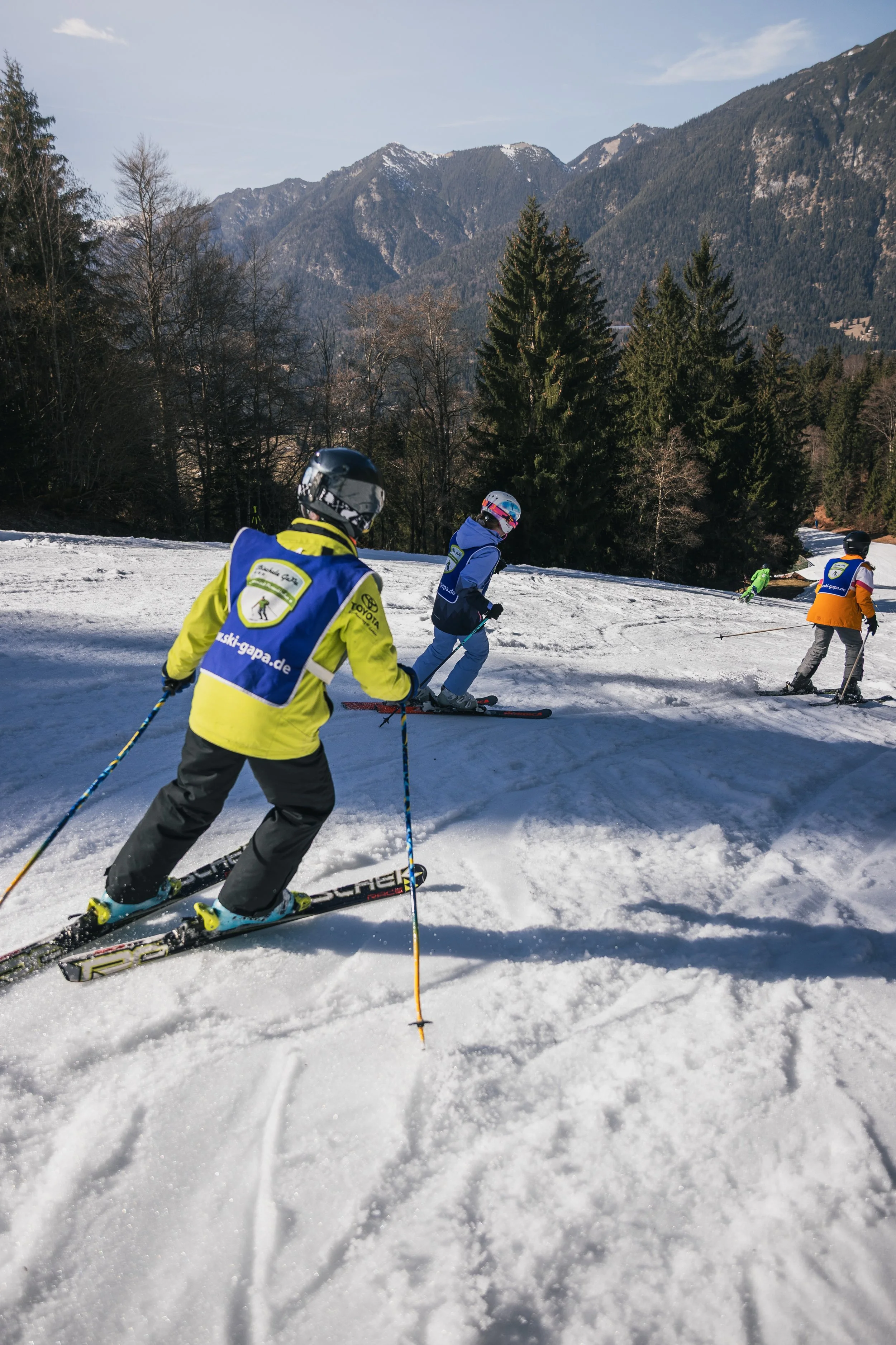 Children skiing down a snow-covered slope surrounded by trees and mountains in the background.