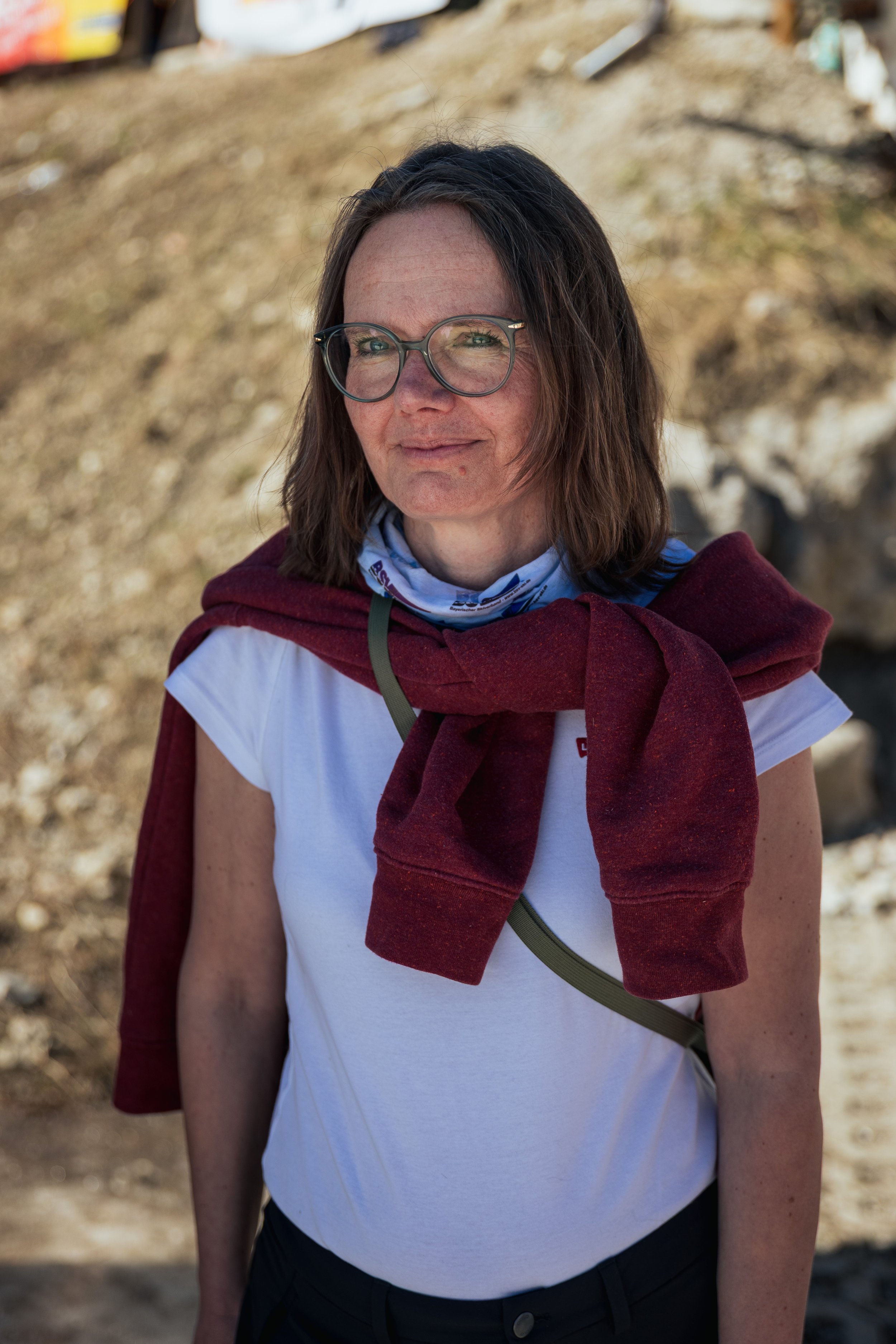 A woman with shoulder-length brown hair and glasses standing outdoors in a rocky area, wearing a white T-shirt with a maroon sweater tied around her shoulders, and a bandana around her neck.