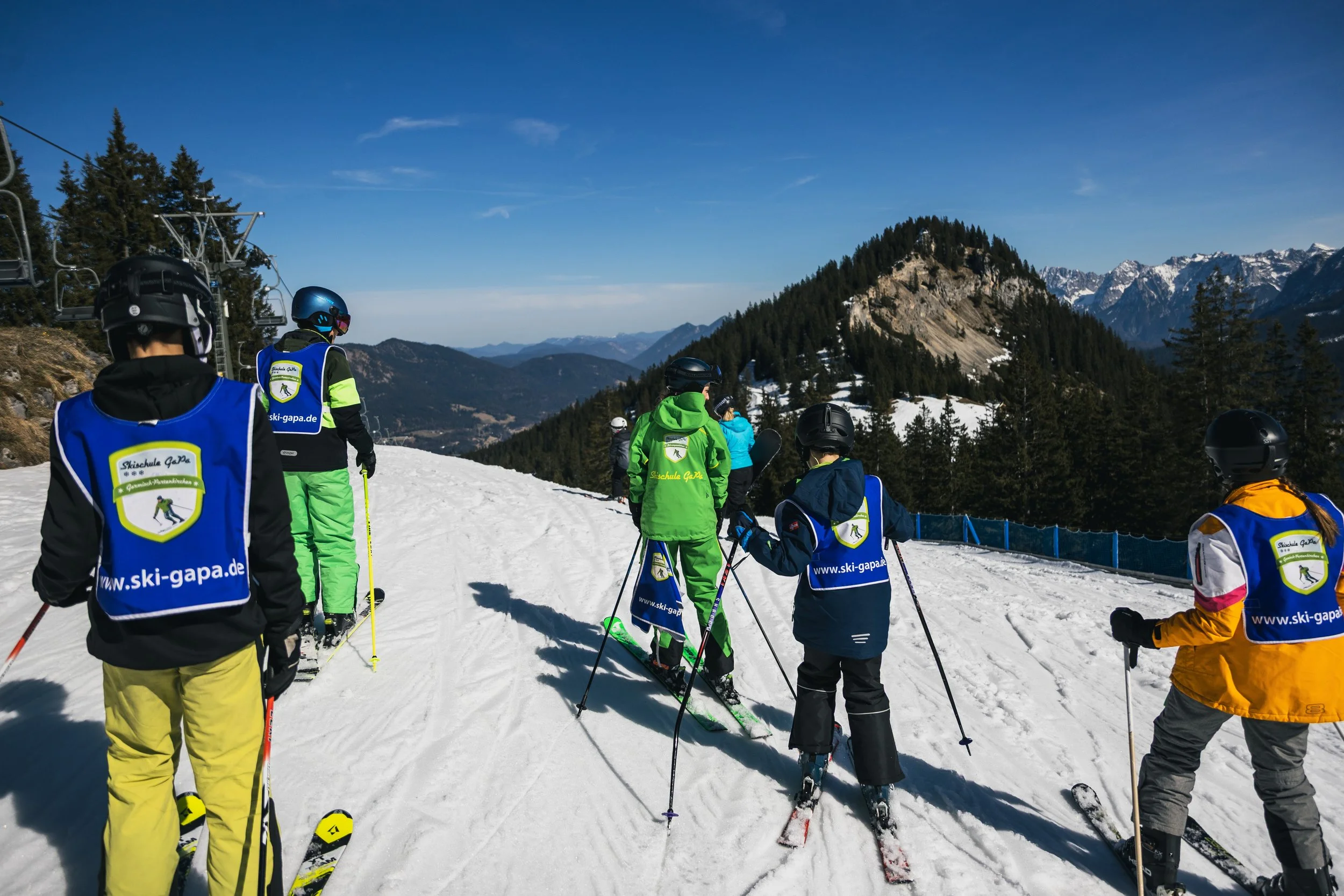 Group of children and adults in colorful ski gear on a snowy mountain slope with pine trees and mountain peaks in the background, preparing for skiing.
