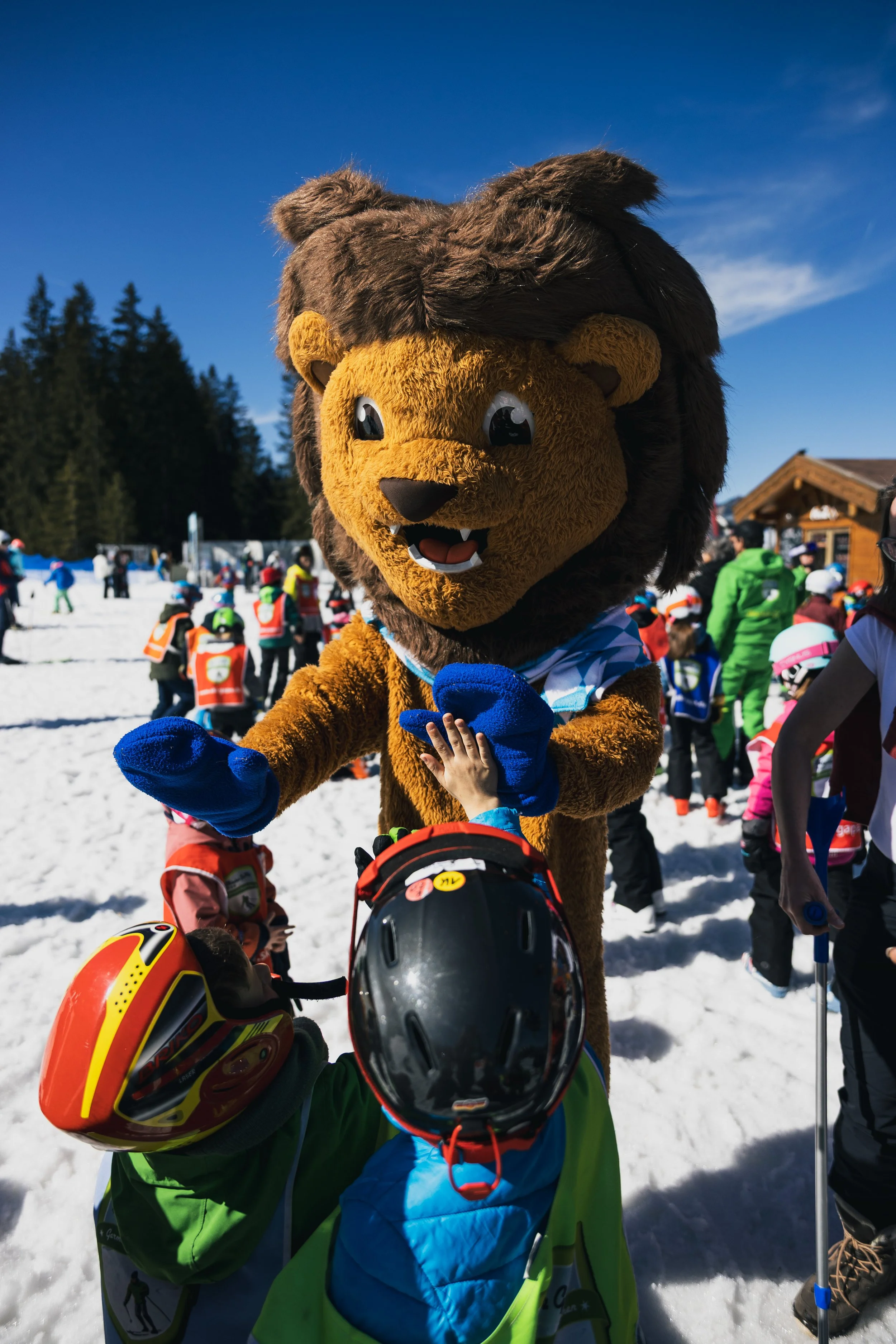 A person in a lion mascot costume high-fives and interacts with children during a snowy outdoor event, with many kids and adults in colorful winter clothing and helmets.