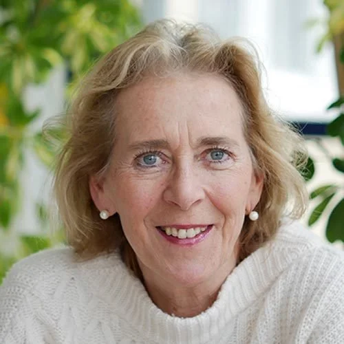 A smiling woman with blonde hair, wearing a white sweater and pearl earrings, in a bright room with green plants in the background.