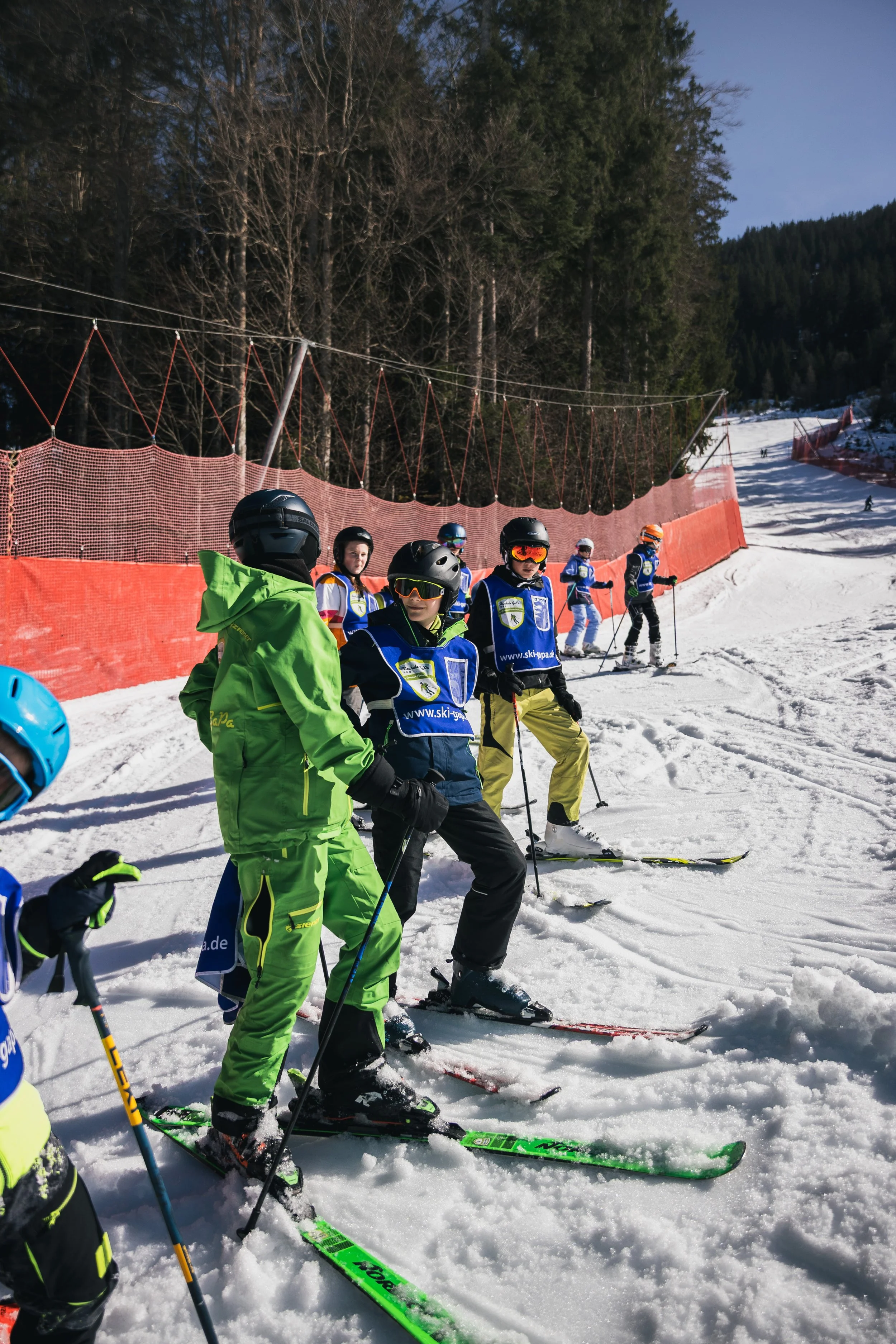 Children in ski gear standing and skiing on a snowy slope at a ski resort, with a red safety net and forested mountains in the background.