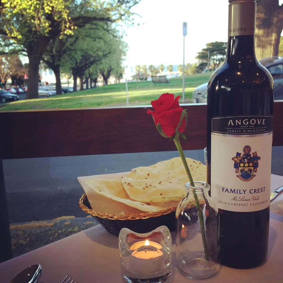 Table setting at Eastern Spice Geelong featuring Angove Family Crest 2014 Cabernet Sauvignon, a lit candle, a red rose in a vase, and a basket of flatbread chips, with green trees and a park visible through the window, highlighting a refined Indian d