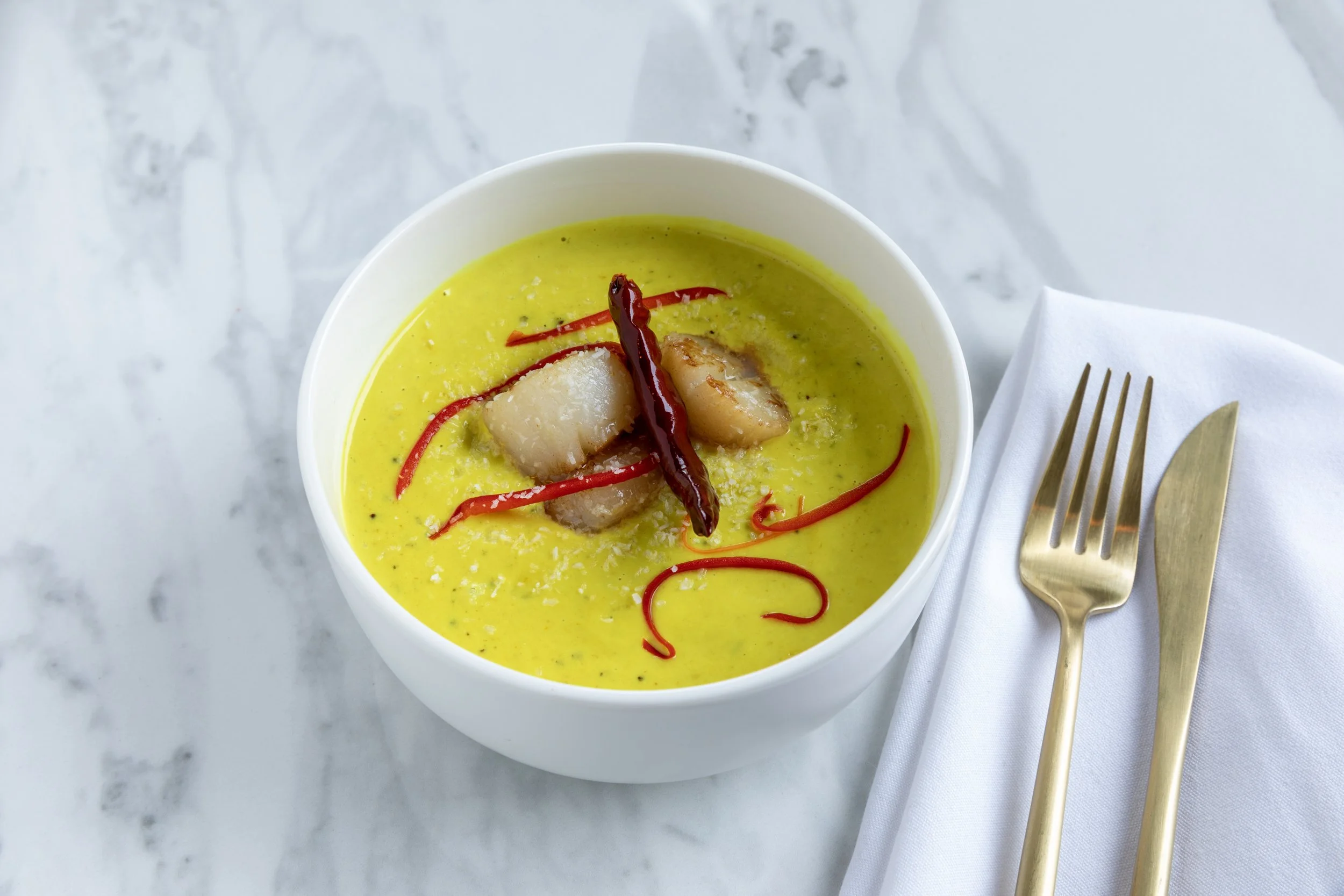 A white bowl filled with bright yellow curry topped with three pieces of scallops, a red chili pepper, and thin strips of red chili.
