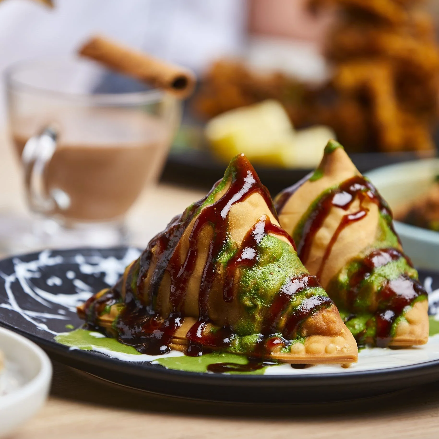Close-up of two samosas topped with chutneys on a black plate, with a cup of chocolate milk in the background.