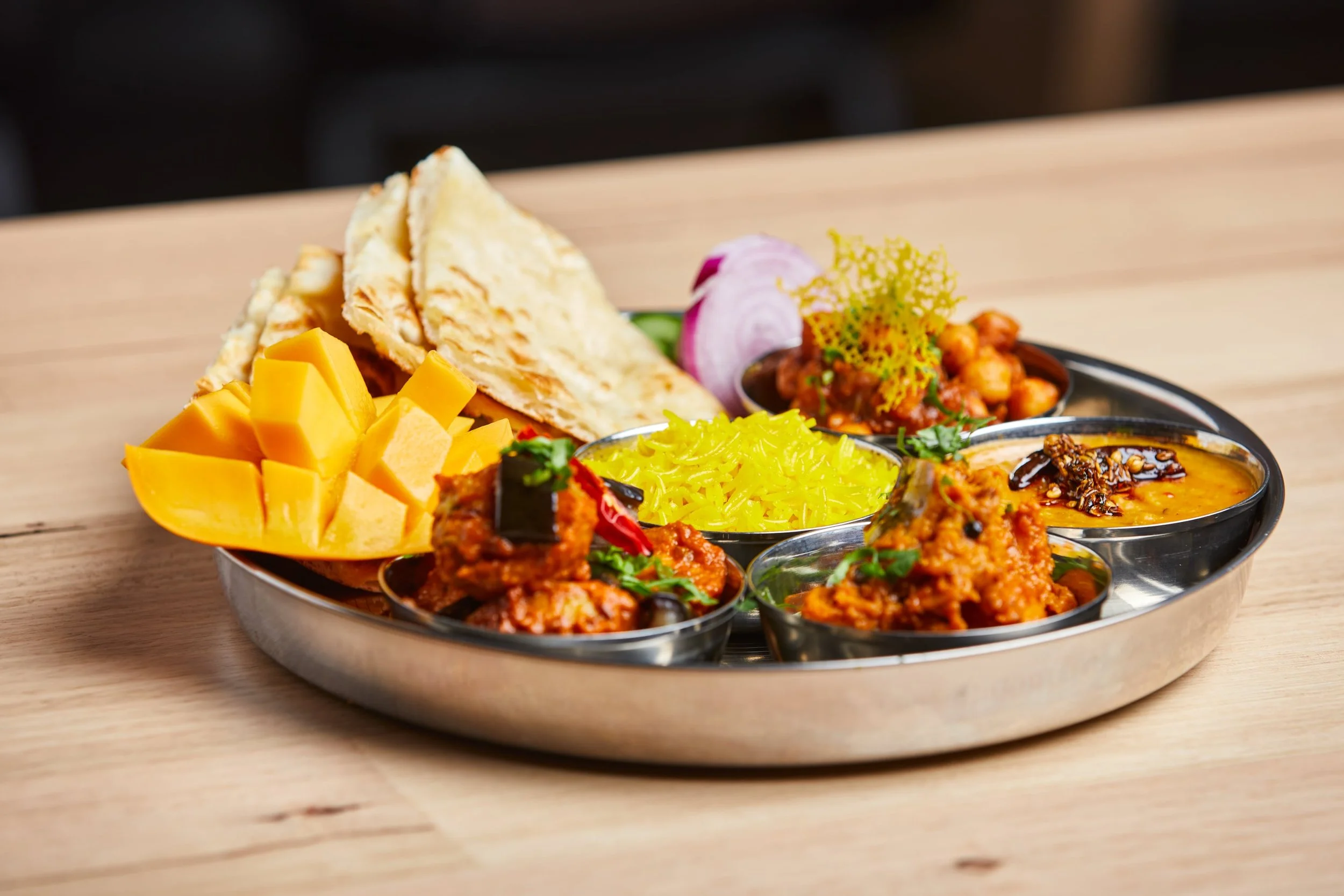 Thali platter with cheese, flatbread, chickpea curry, vegetable curry, rice, onions, and garnishes on a wooden table.