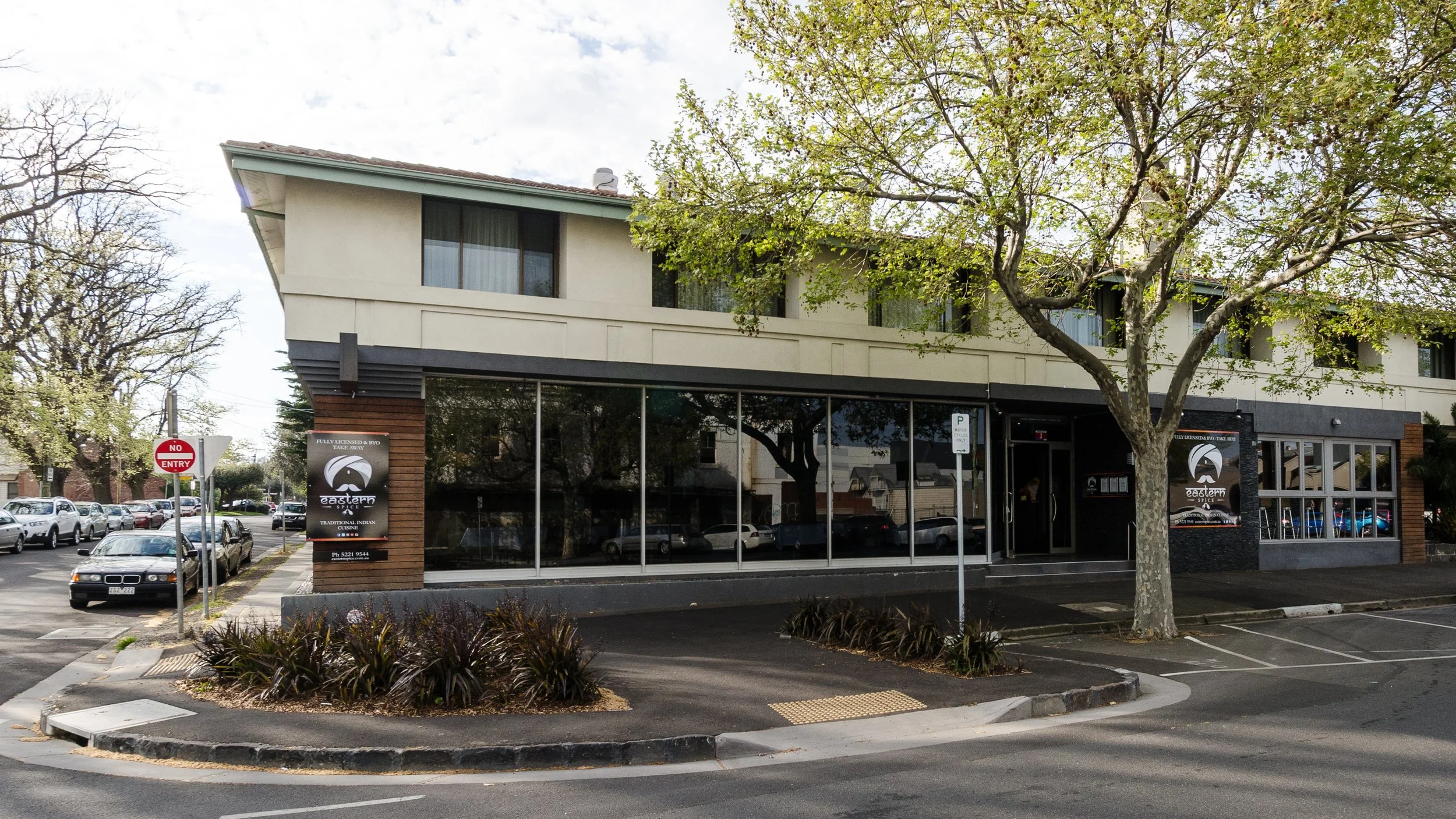Street view of a two-story building with a restaurant called Eastern, a parking lot in front, and a tree partially shading the building.