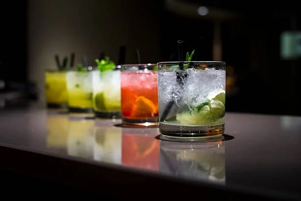 Five colorful cocktails with ice and garnishes arranged in a row on a reflective bar counter.