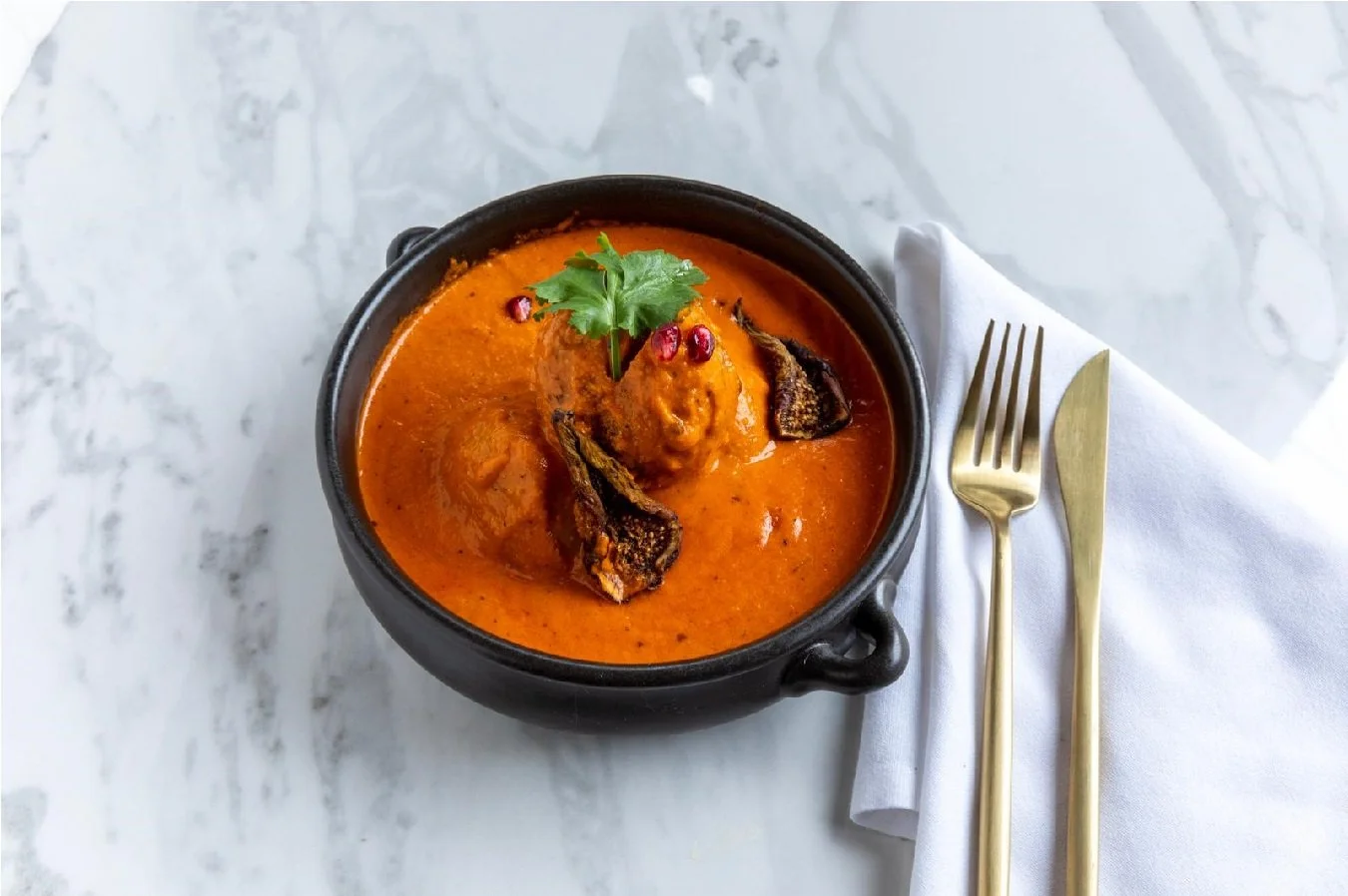 A black bowl of Indian butter chicken curry garnished with cilantro and pomegranate seeds, served alongside a white napkin with a gold fork and knife on a marble surface.