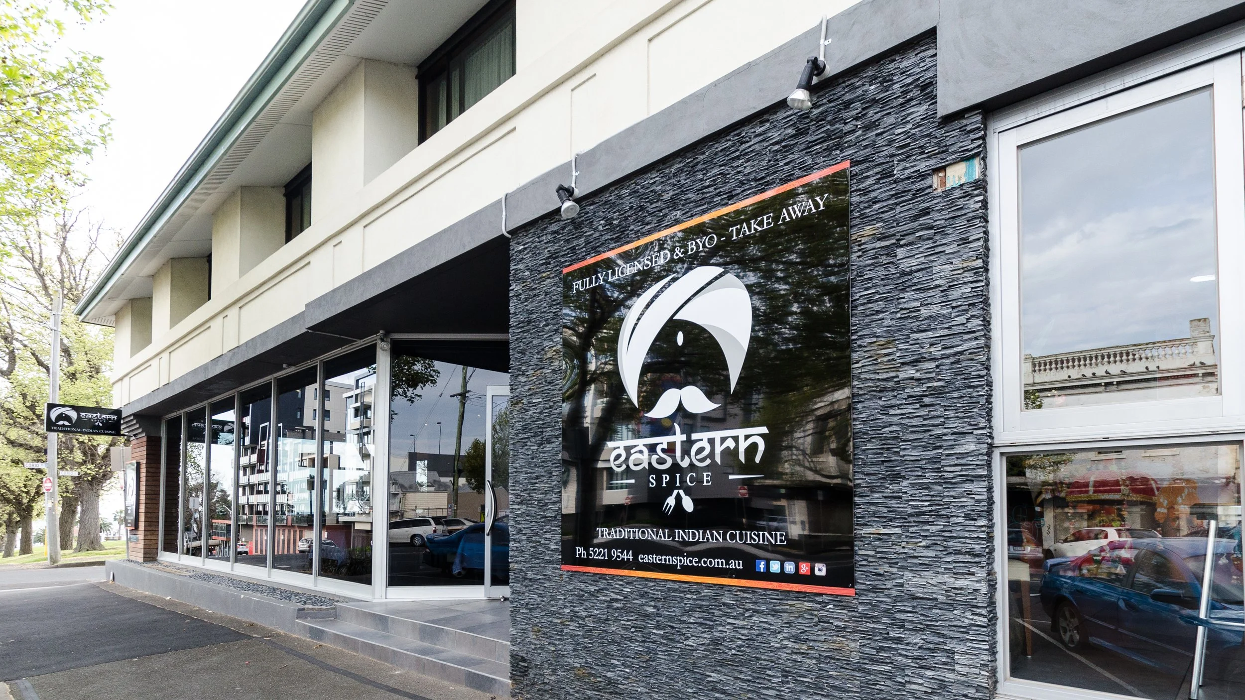 Exterior view of Eastern Spice, a restaurant specializing in traditional Indian cuisine, with signage on a textured wall and large windows reflecting parked cars and trees outside.