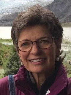 Woman with glasses, smiling, in front of trees and water.