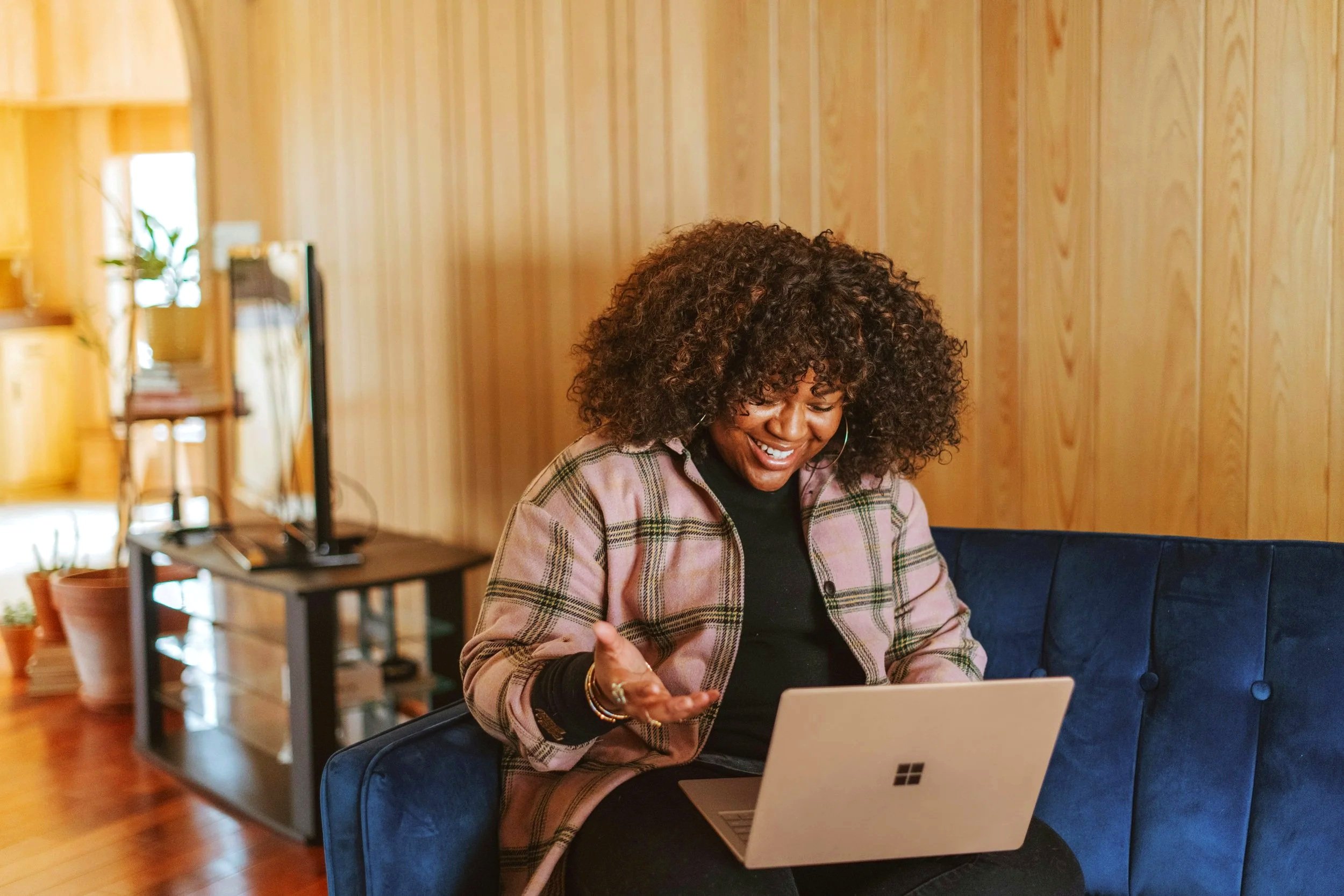 A woman with curly hair smiling while working on a laptop in a cozy wooden-paneled room.