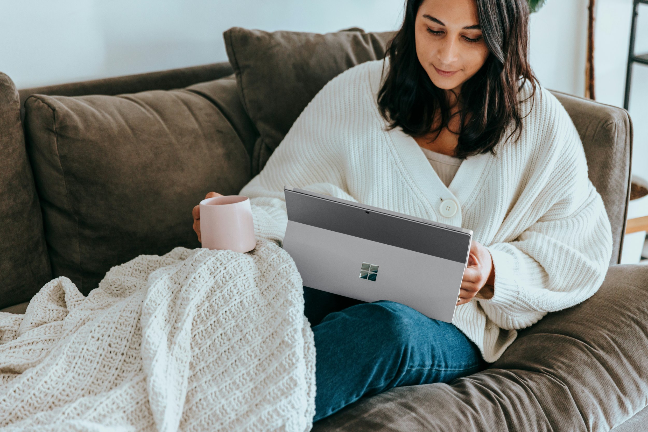 A woman with dark hair wearing a white cardigan sitting on a brown couch, looking at a device in her hands, with a white mug and a chunky cream-colored blanket nearby.