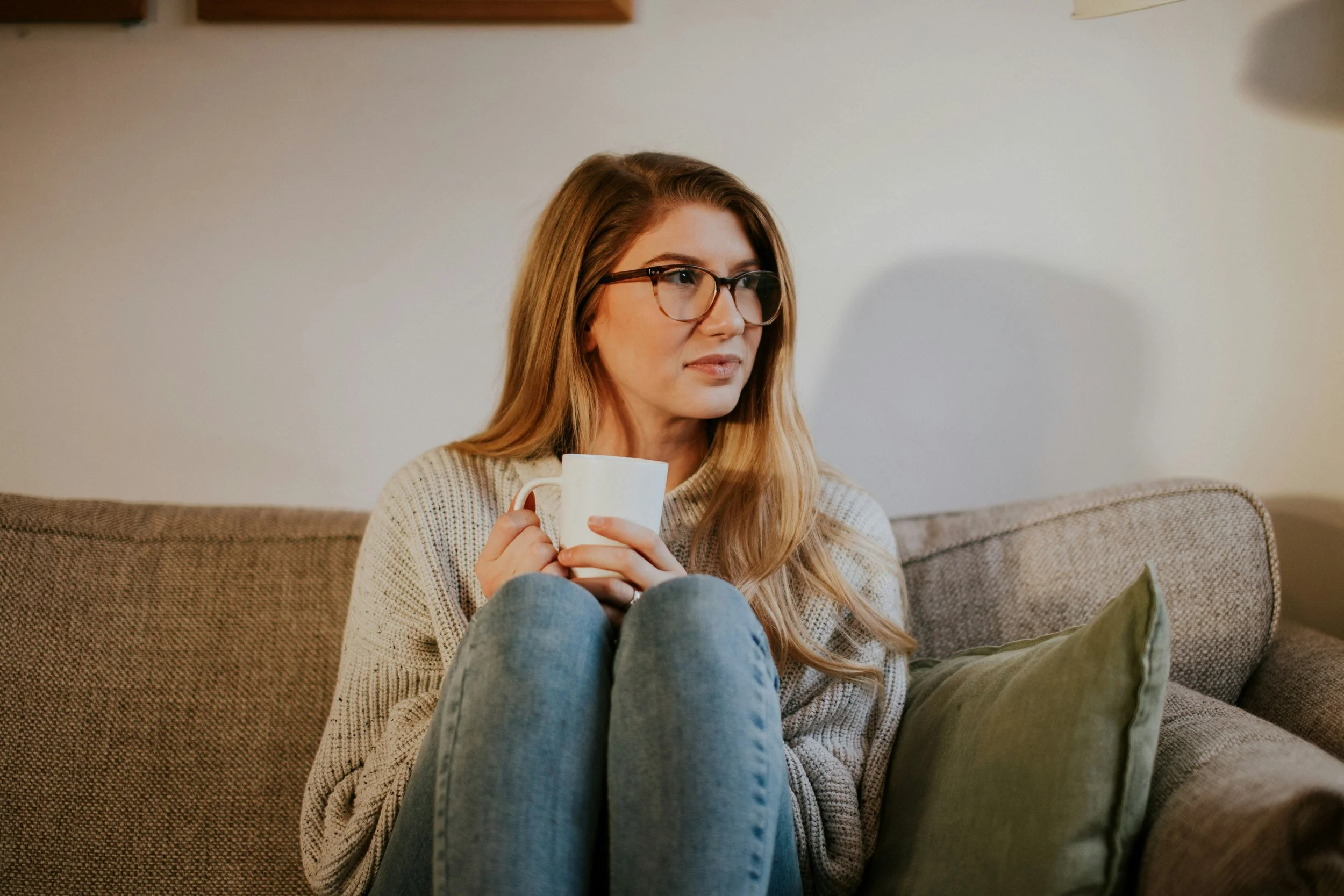A woman with long blonde hair and glasses sitting on a brown couch with a white mug in her hands.