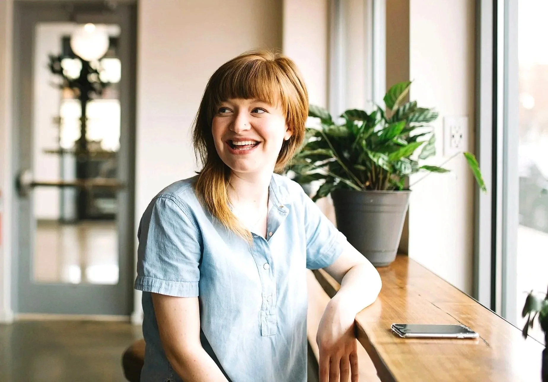 A woman with red hair and a light blue denim shirt sitting on a stool at a wooden counter, smiling and looking to her left, next to a potted plant and a smartphone, inside a cafe with natural light.