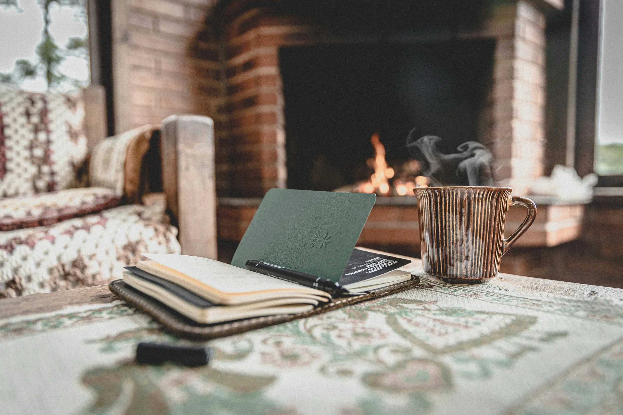 A table with books, a notebook, a pen, a mug with steam, and a green card, in front of a fireplace and window.
