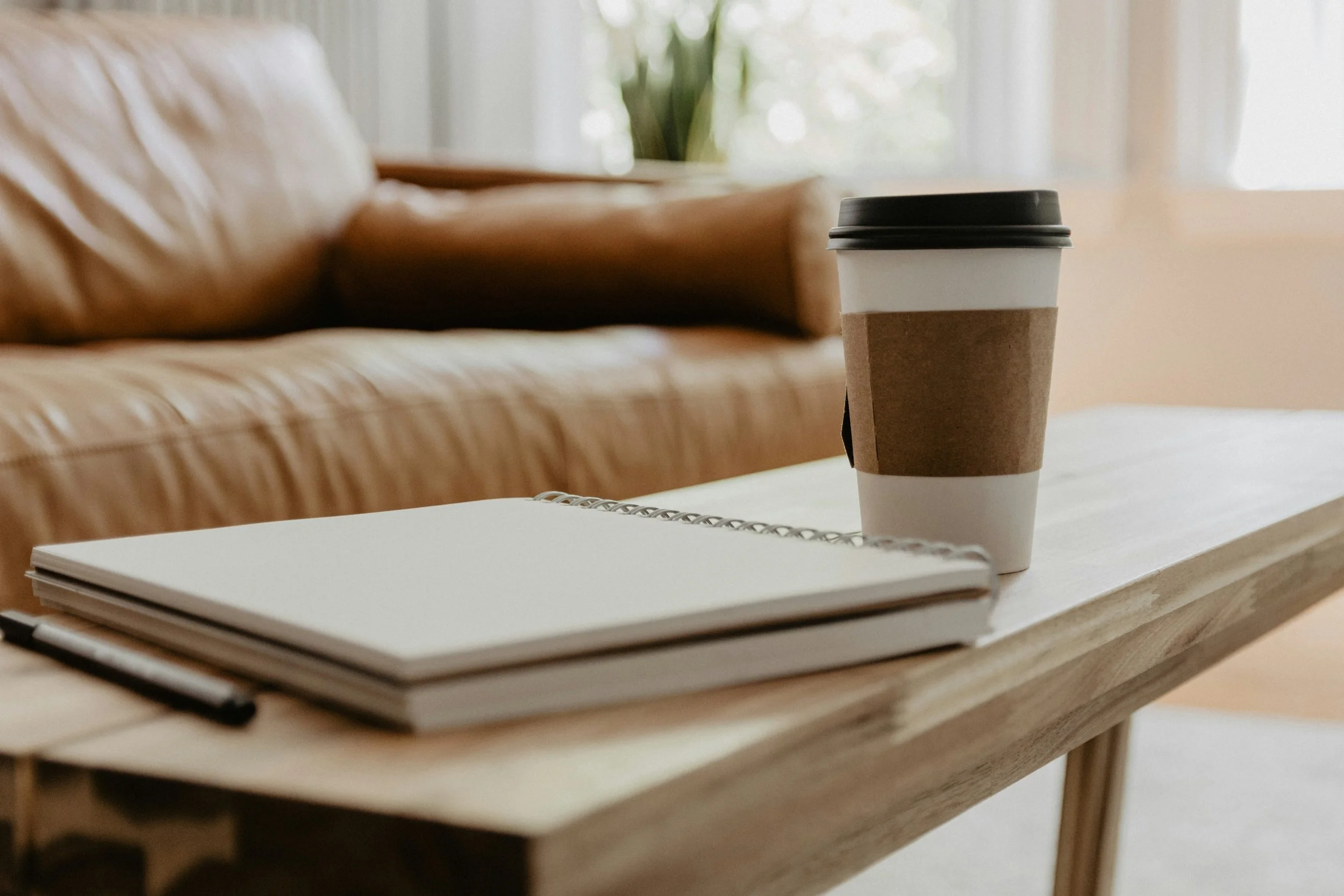 A coffee cup with a black lid and a cardboard sleeve on a white surface. An open notebook with a spiral binding is in the foreground, and a blurred background shows a couch with a cushion and a window with curtains.