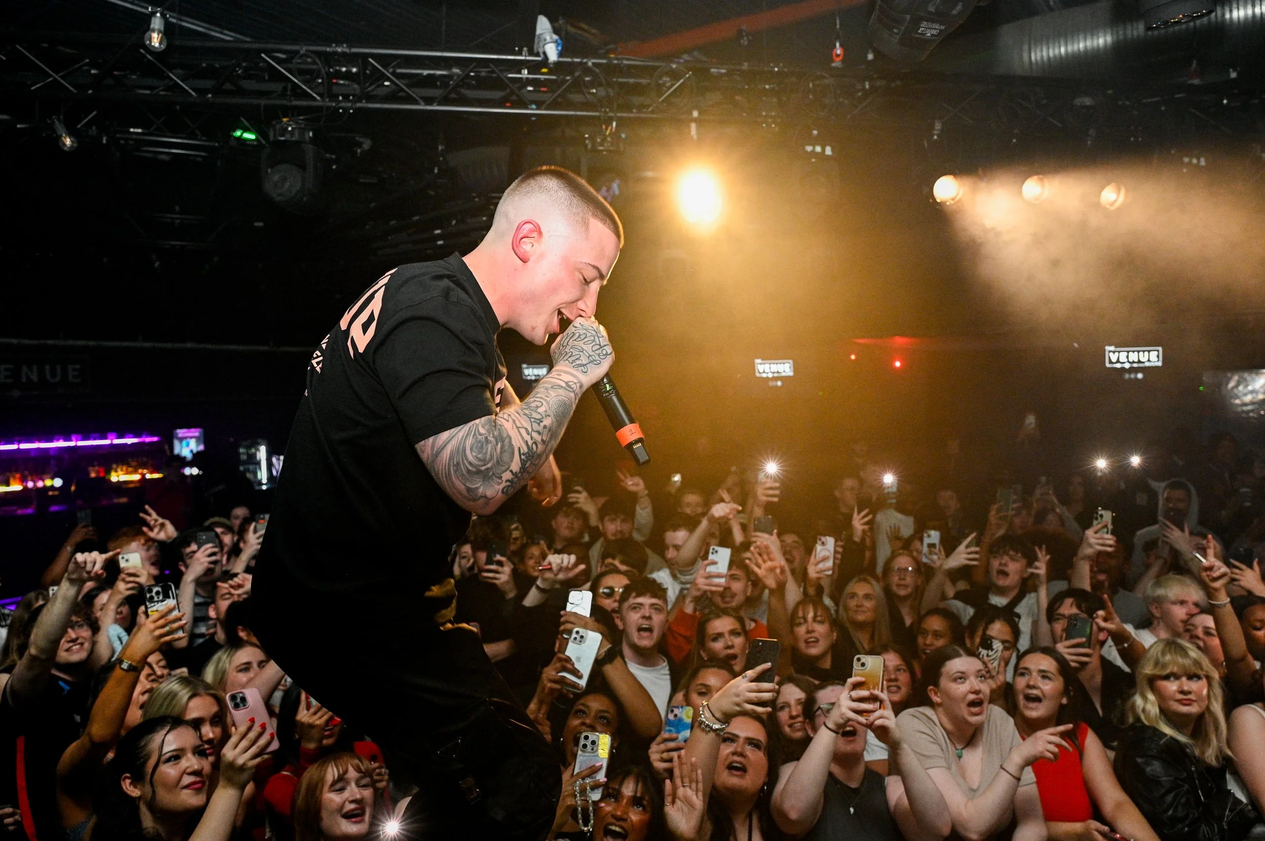 Performer singing into a microphone at a concert while crowd members take photos and cheer.