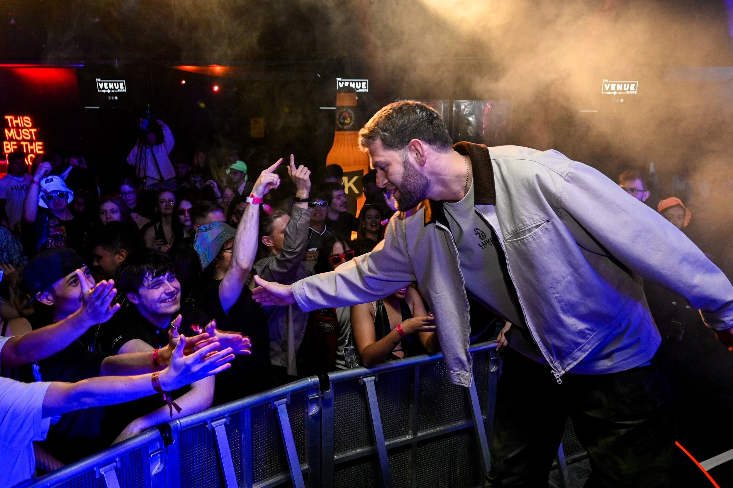 A male performer greeting and shaking hands with fans at a concert or event, with a crowd of young excited people behind a barrier.