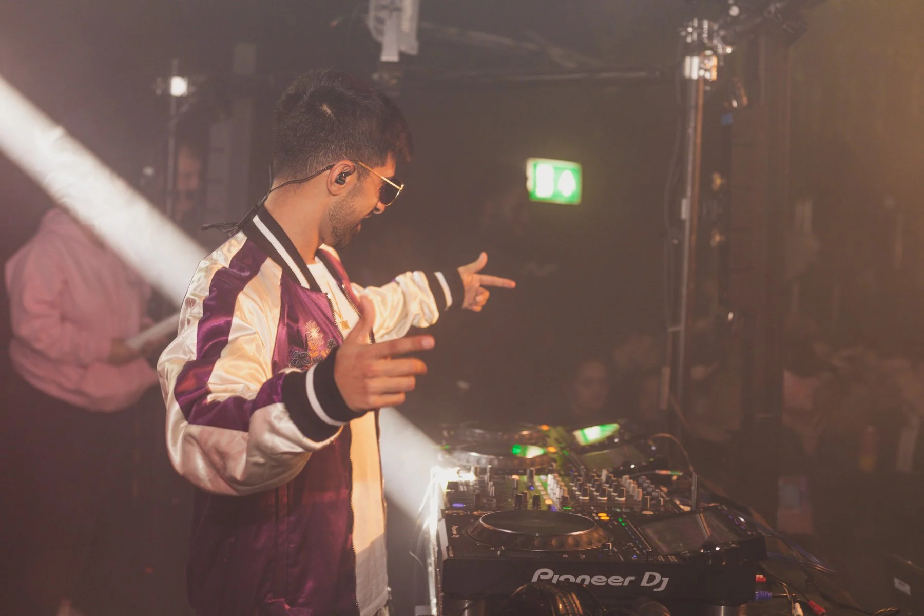 DJ wearing sunglasses and a satin jacket, dancing at a DJ booth with Pioneer DJ equipment, in a dimly lit nightclub.