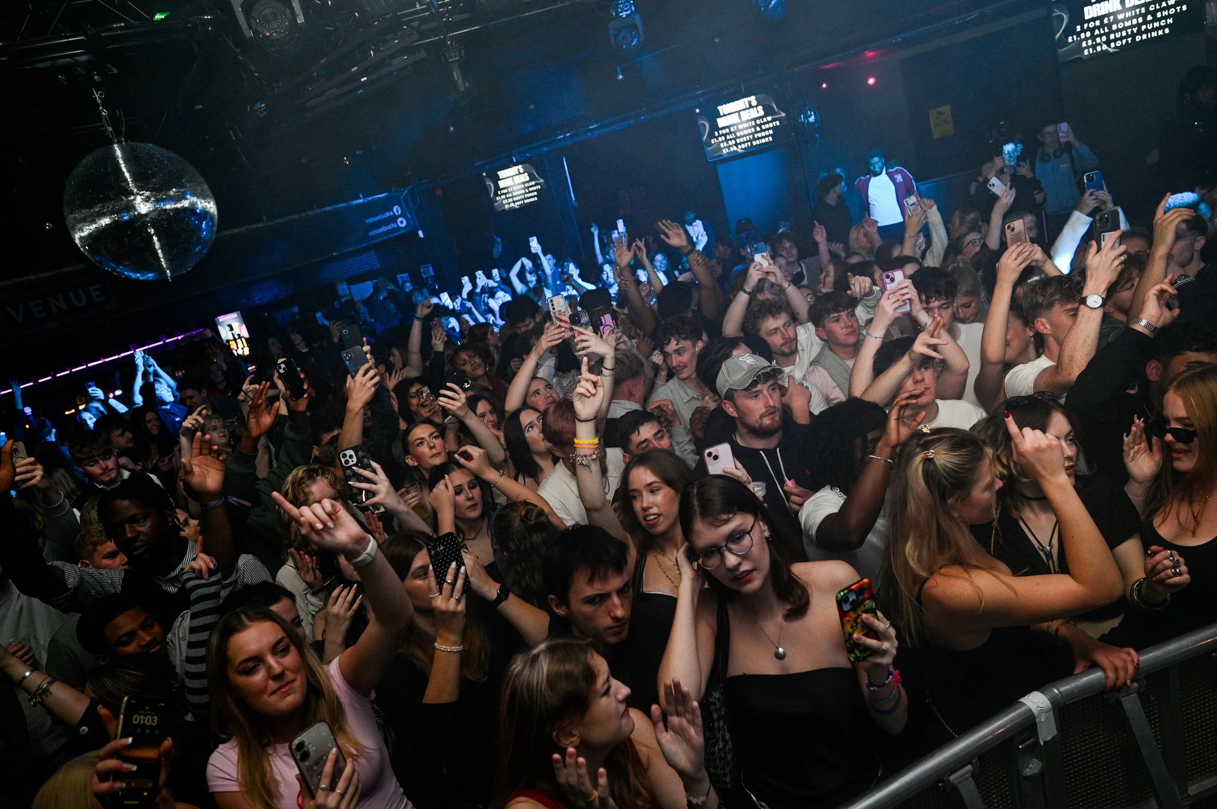 Crowd of young people at a concert or nightclub, many holding up phones to record in a dark, blue-lit environment with a disco ball overhead.