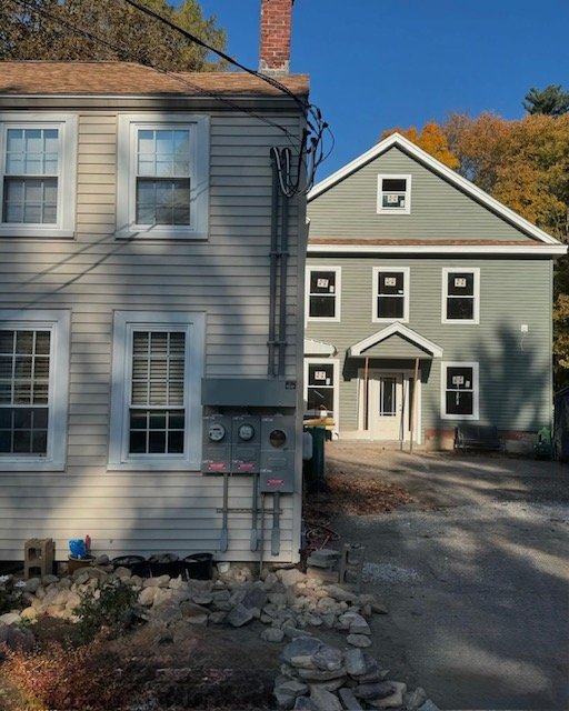 Two adjacent residential houses with light-colored siding, surrounded by trees. The foreground house is off-white, with visible windows and utility meters on the side. The background house is a soft green with a small porch and multiple windows, situ