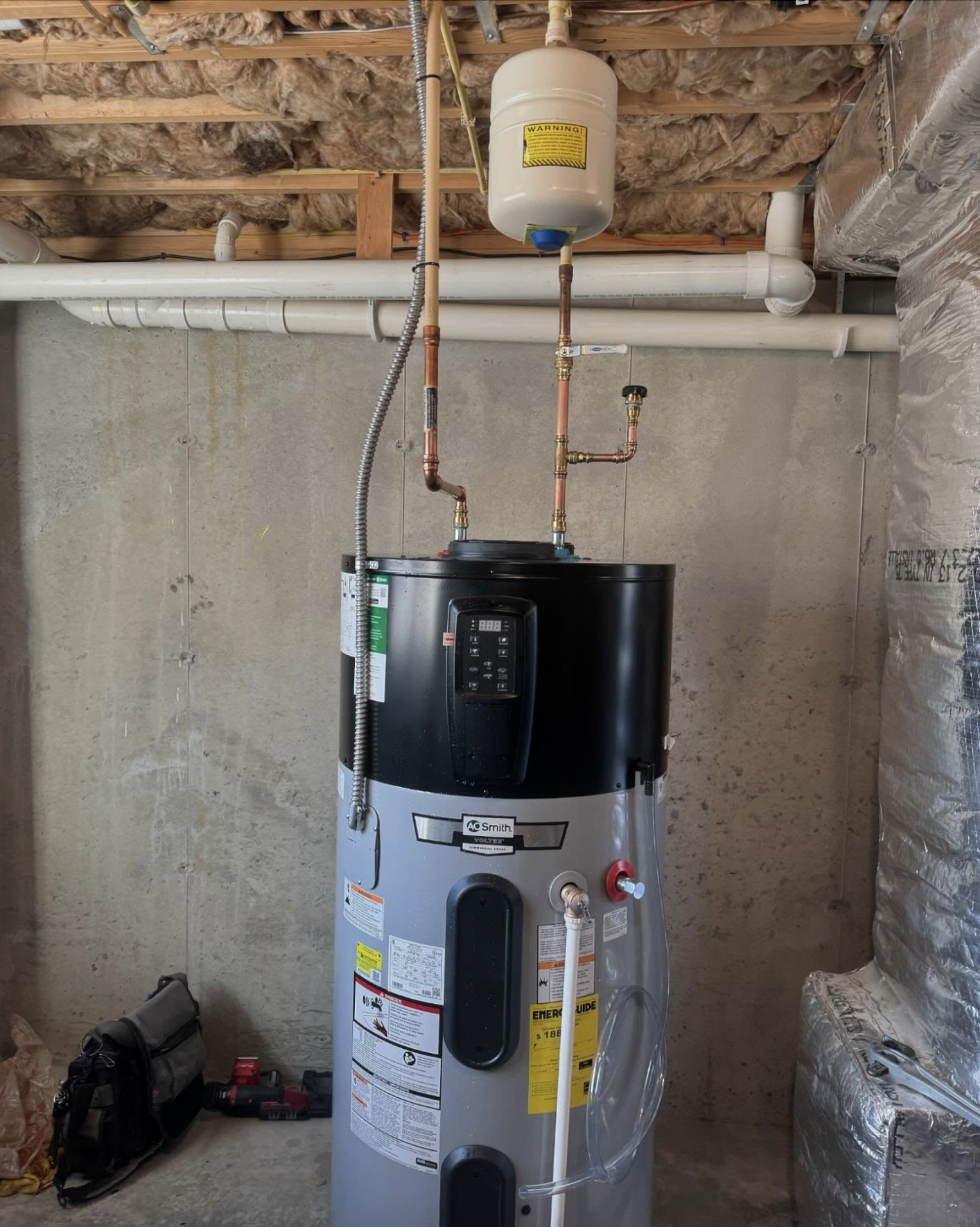 A water heater installed in a basement with pipes and an expansion tank connected above.