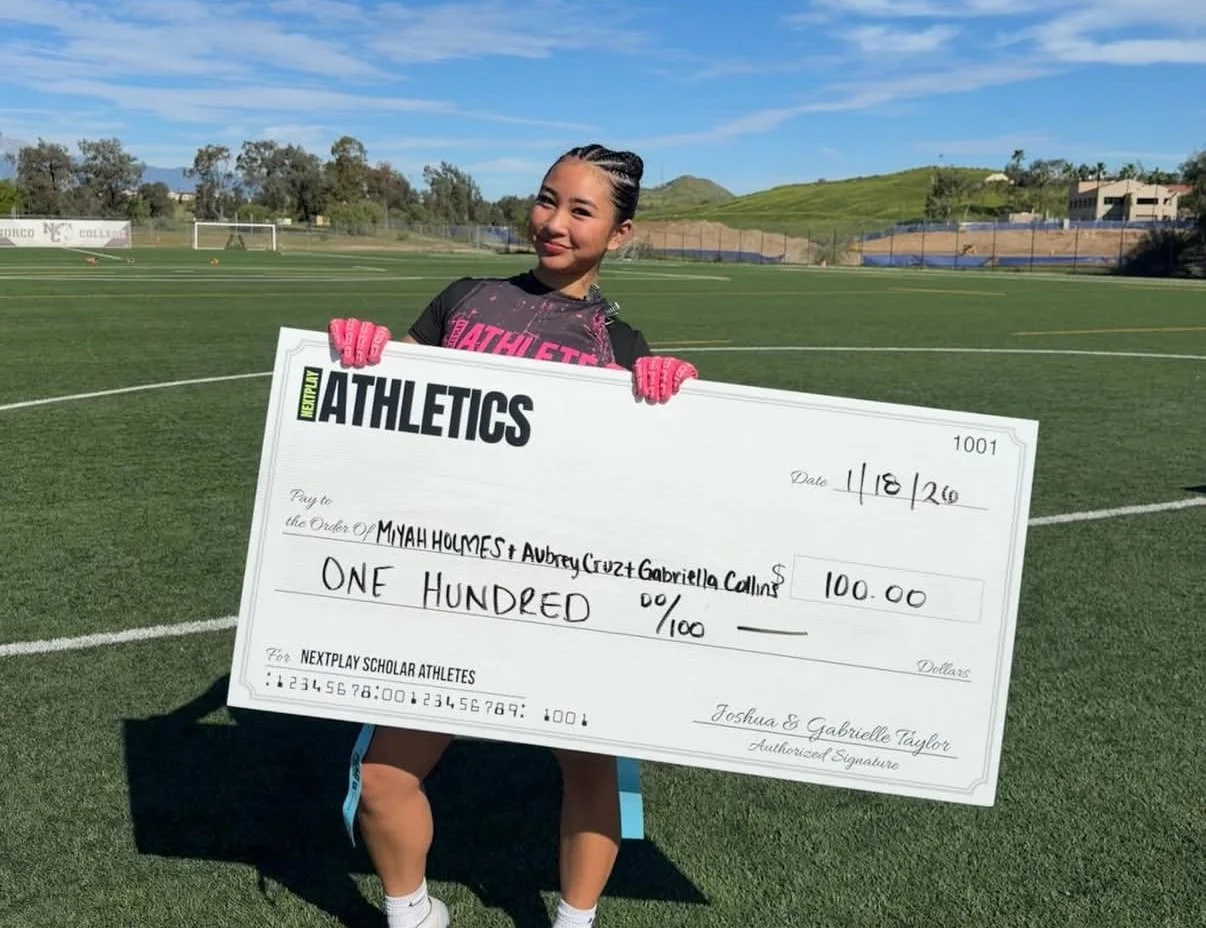Young girl on a sports field holding a large check for 100 dollars made out to MYAH HOMES and Aubrey Cruz and Gabriella Collins, dated 4/18/20, with a bright smile, wearing sports attire and pink gloves, with a clear sky overhead.