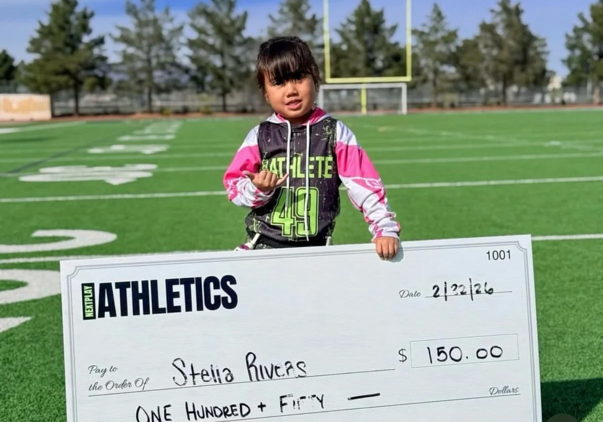 A young girl standing on a football field holding an oversized check for $150 made out to Stella Rivers, with trees and a goalpost in the background.