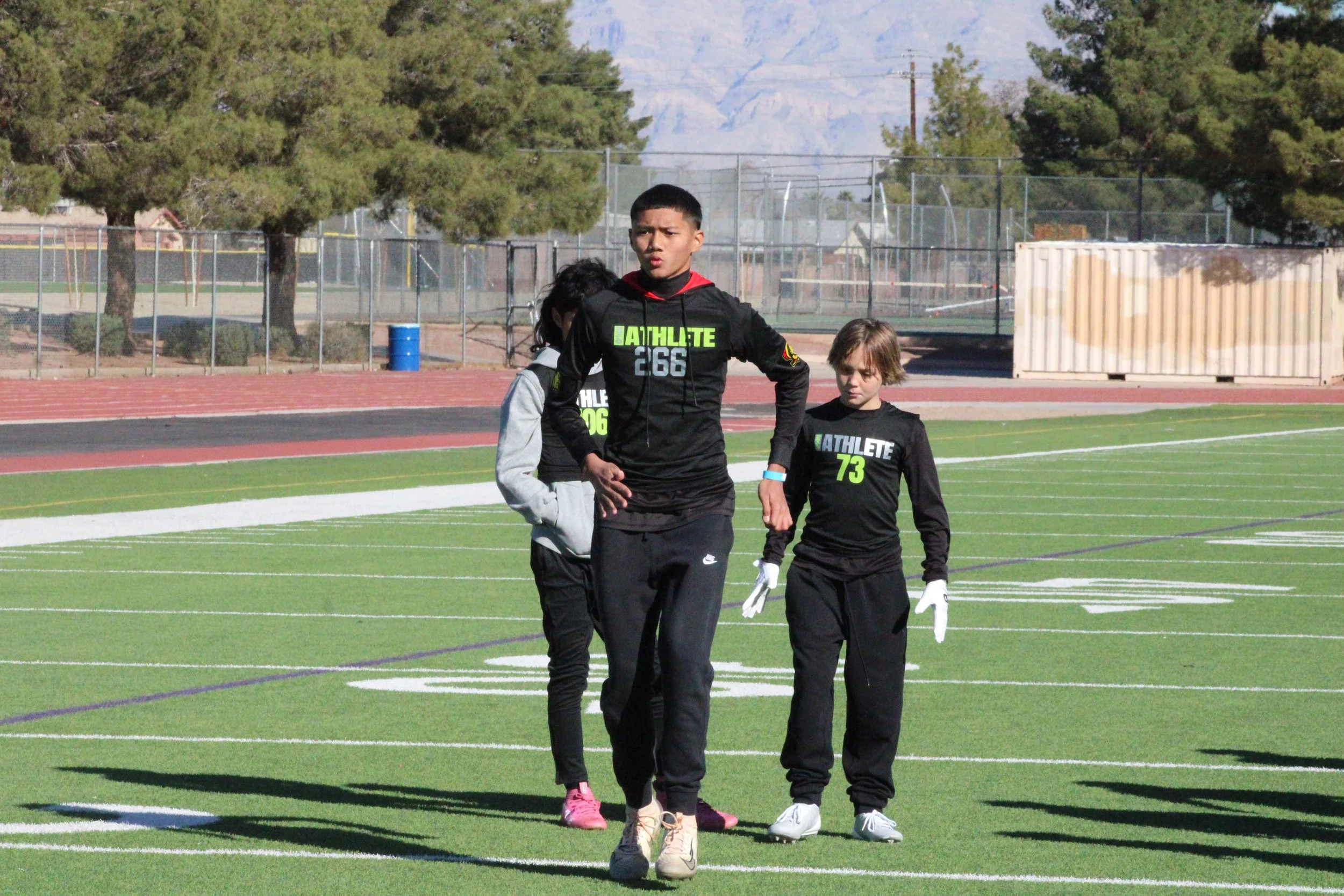 Three young athletes on a sports field, wearing black uniforms with neon green and white lettering, with trees and a fence in the background.