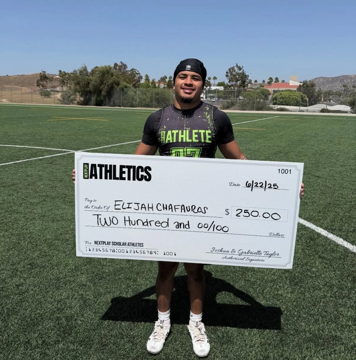 Young man in athletic attire holding a large check on a football field, with a blue sky and trees in the background.