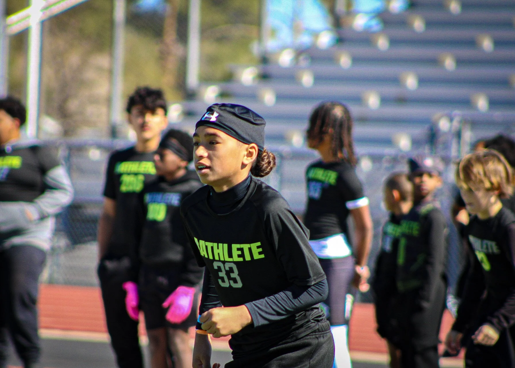 A group of young athletes, mostly girls, on a track field during a sports event, wearing black jerseys with the word 'ATHLETE' and numbers, some with gloves and headbands.