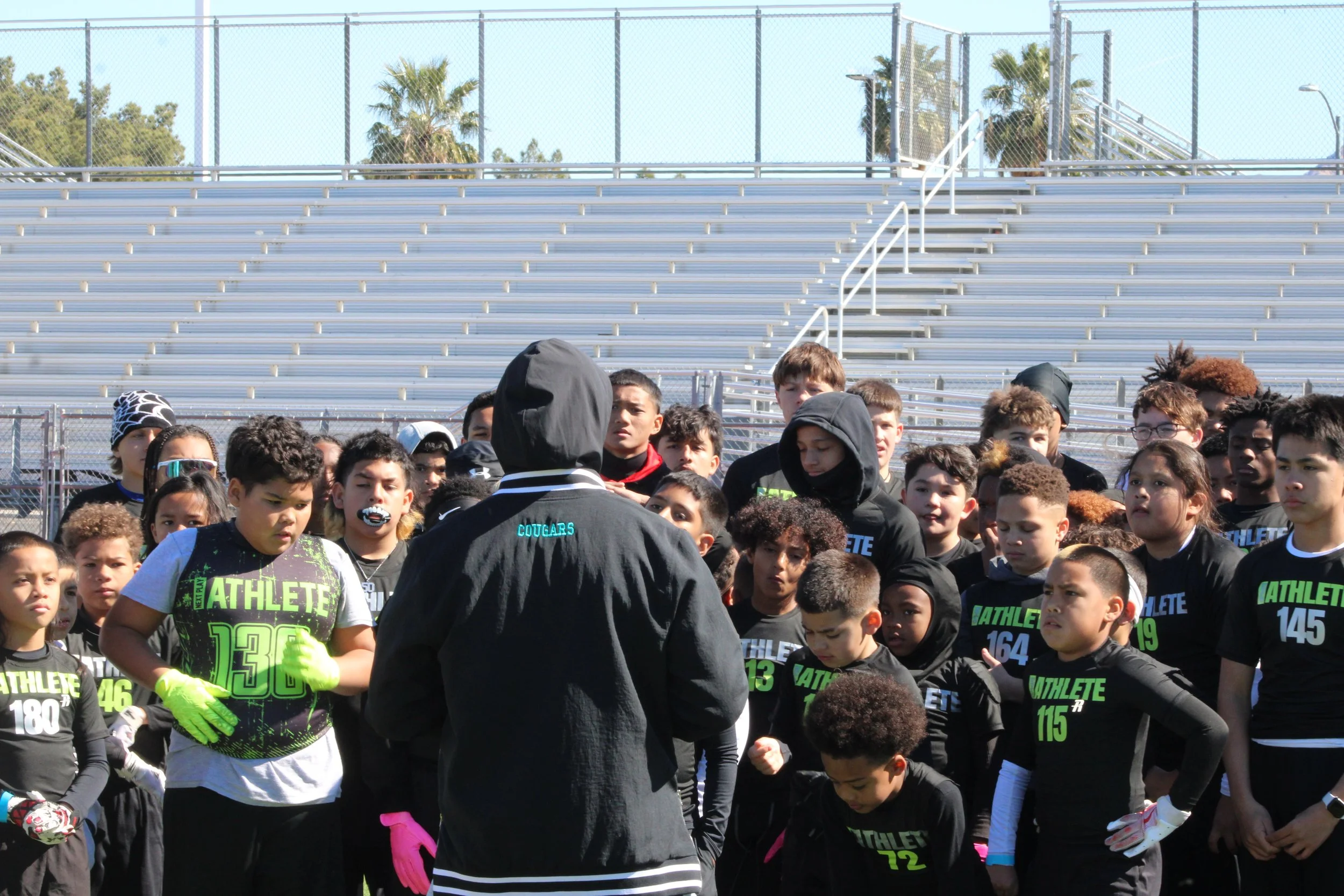 Young athletes and their coach assembled on a football field for a team meeting or instructions, with stadium bleachers and palm trees in the background.