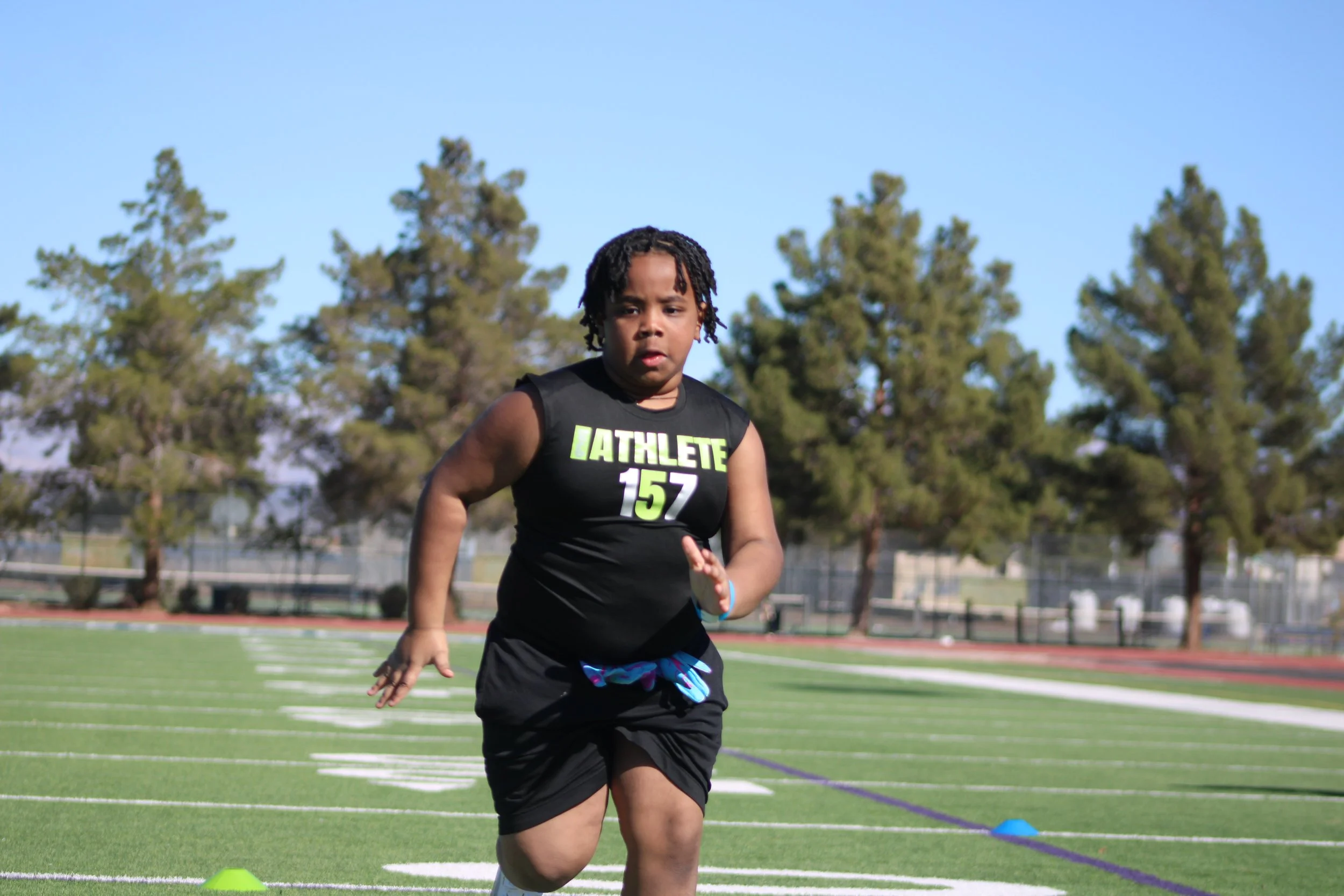 Young boy wearing a black athletic shirt with 'LATHLETE 157', running on a track with trees and clear blue sky in the background.