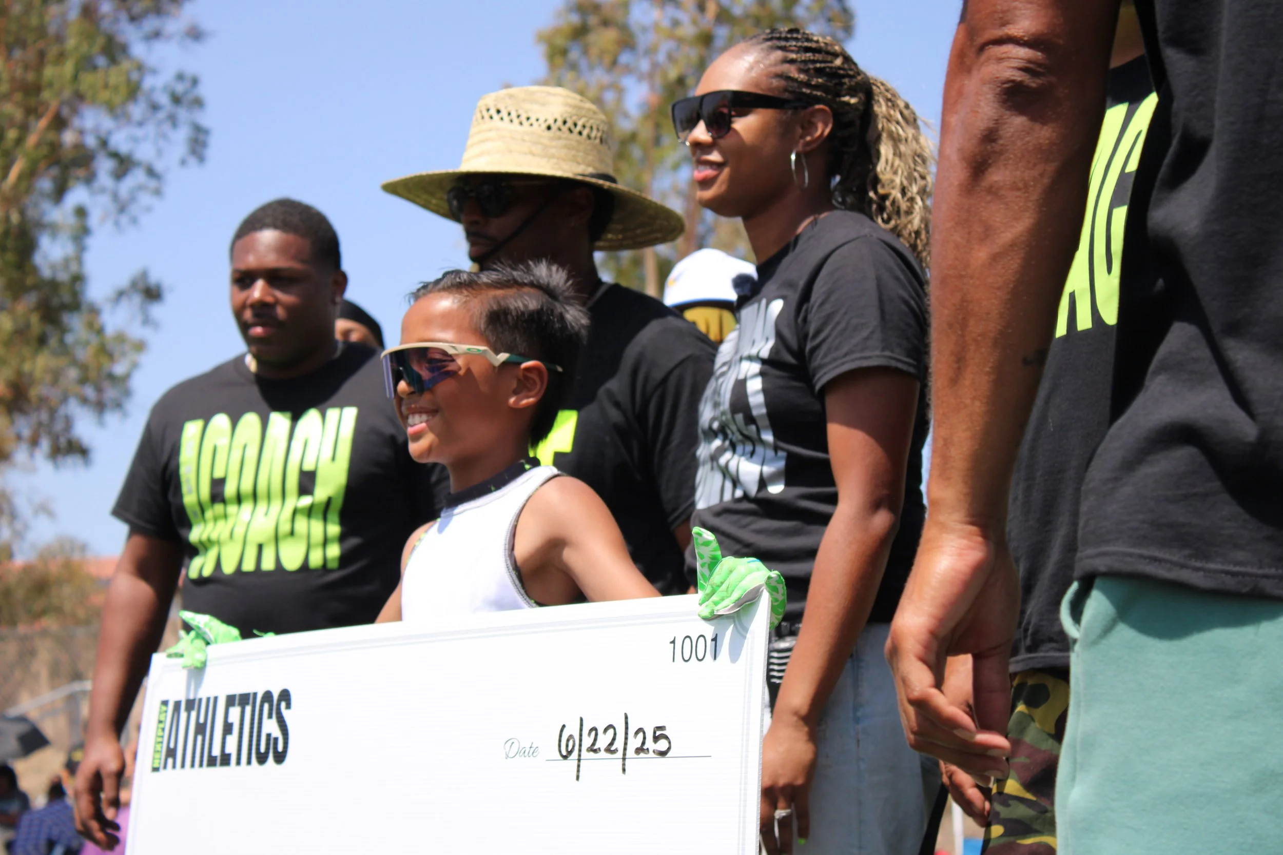 A group of six people standing outdoors, with a young boy in the center holding a large check dated 6/22/25, surrounded by adults wearing black shirts with bright green text. The children and adults are smiling and wearing sunglasses, with a clear sky and trees in the background.