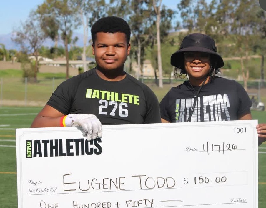 Young man and woman standing outdoors on a sports field, holding a large check made out to Eugene Todd for $150, with trees and a fence in the background.