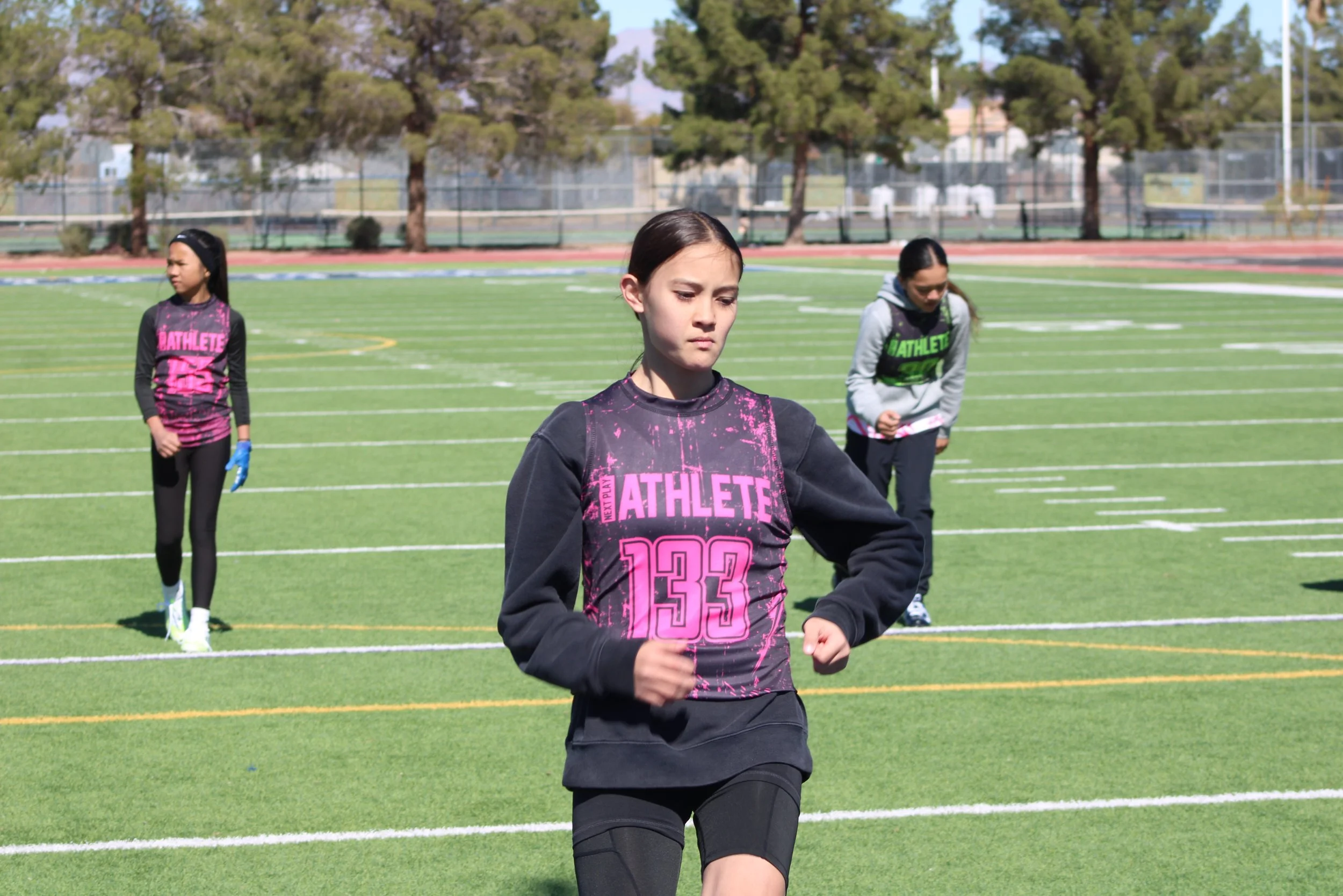 Young female athletes jogging on a green field at a sports stadium, with trees and a fence in the background.