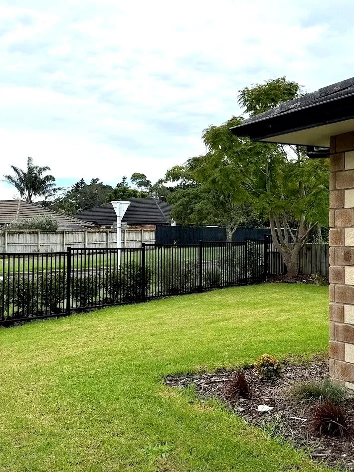 A backyard with a green lawn, a black metal fence, trees, and a house with brick and dark roofing. Overcast sky.
