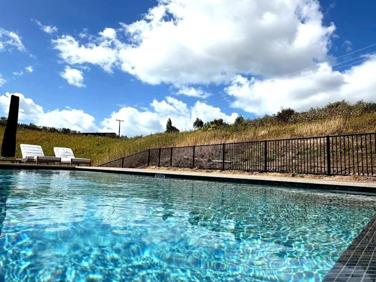 Outdoor swimming pool with clear water, surrounded by a black metal fence and white lounge chairs, on a grassy hill under a partly cloudy sky.