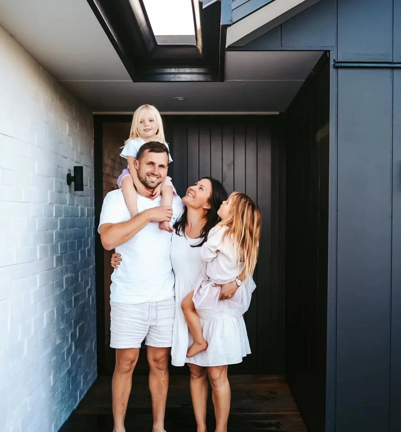 A family of four standing on a porch, smiling and looking at each other, with a man carrying a young girl on his shoulders, and a woman holding another young girl in her arms, against a modern house exterior with dark and white walls.