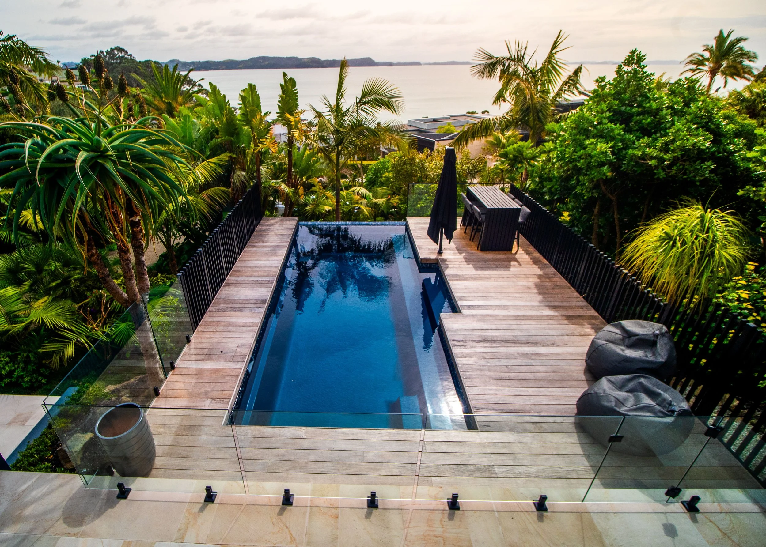 A backyard with a rectangular swimming pool, surrounded by a wooden deck and lush green tropical plants, overlooking a body of water and distant land under a cloudy sky.