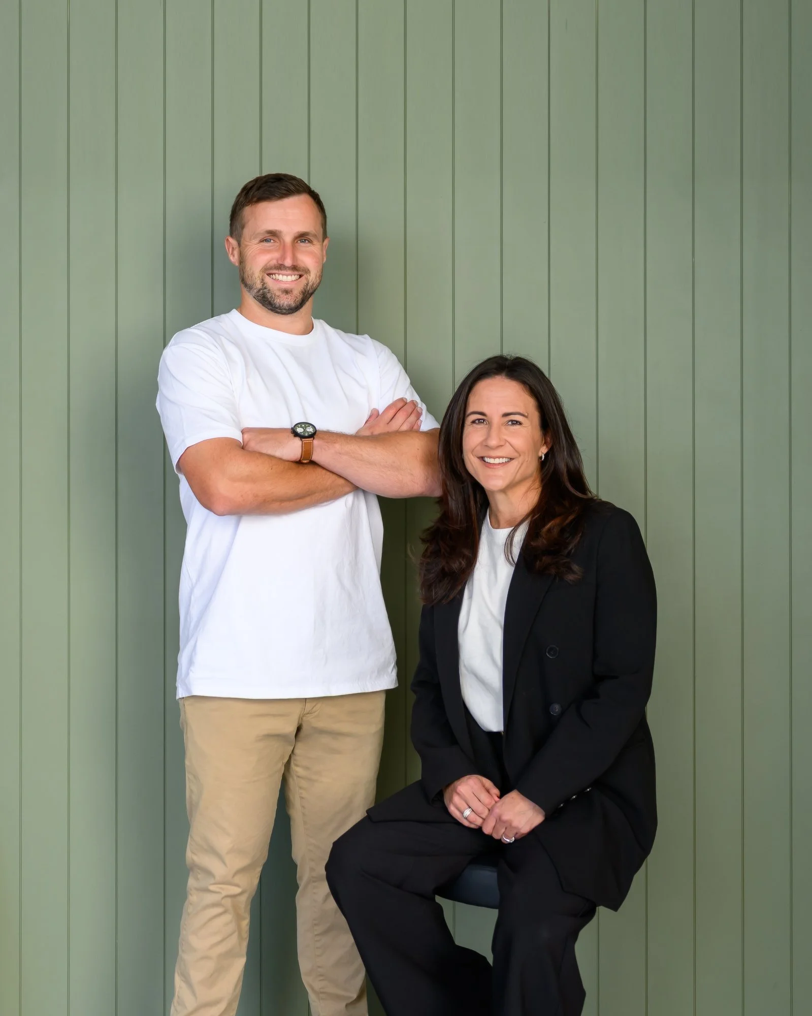 A smiling man standing with arms crossed, wearing a white T-shirt and beige pants, next to a smiling woman sitting on a chair, wearing a black blazer and pants, against a green paneled wall.