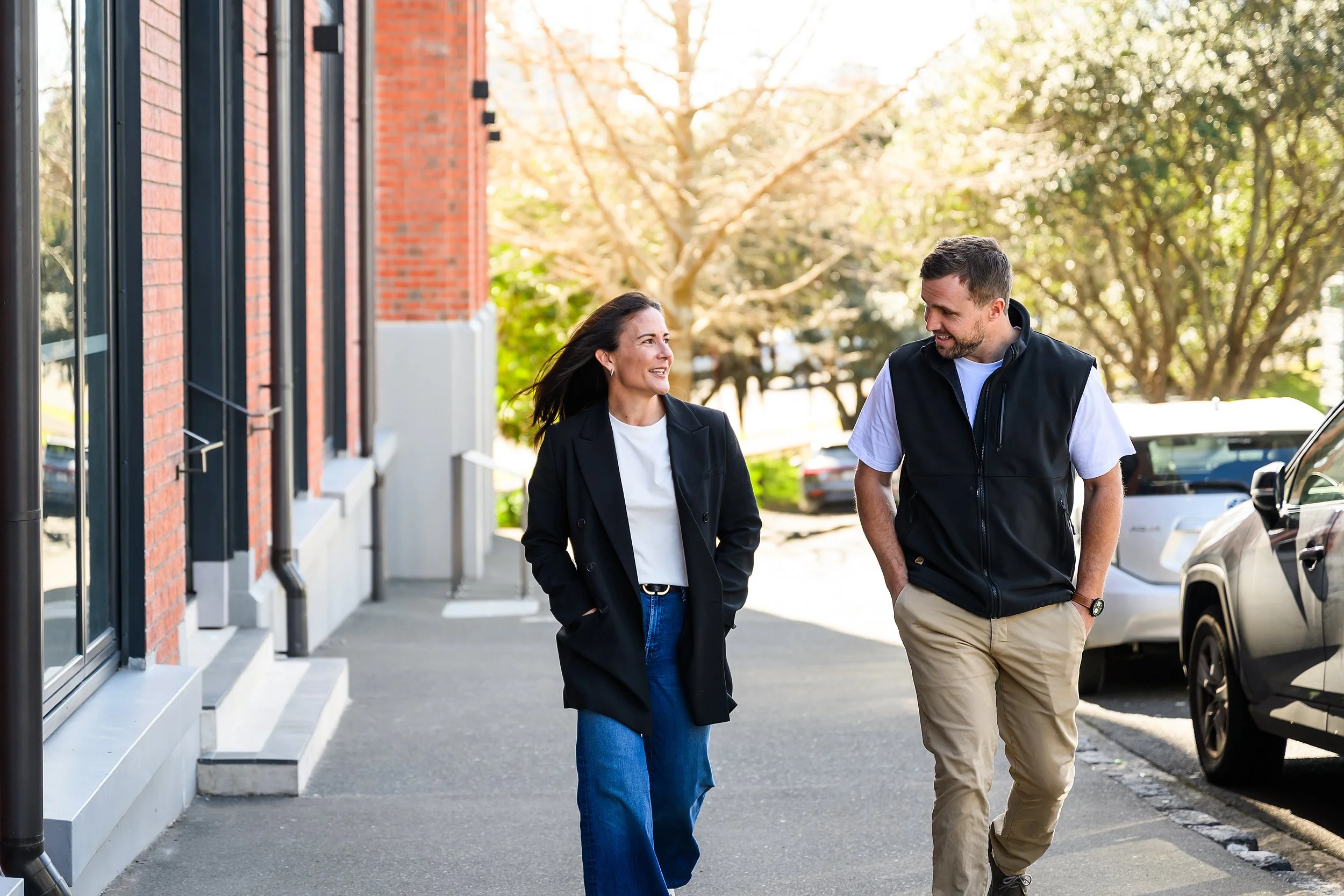 A man and woman walking and talking outside on a sunny day near a red brick building and parked cars.