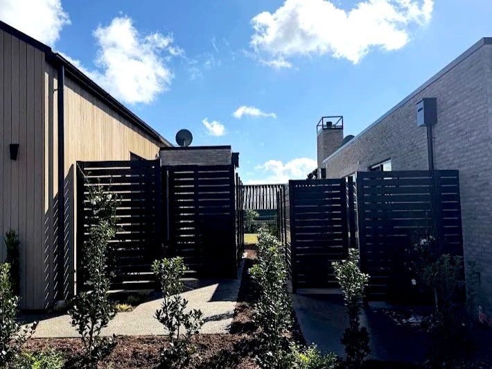 View of a modern outdoor backyard space with black privacy fences, small shrubs, and a pathway between buildings under a blue sky with clouds.