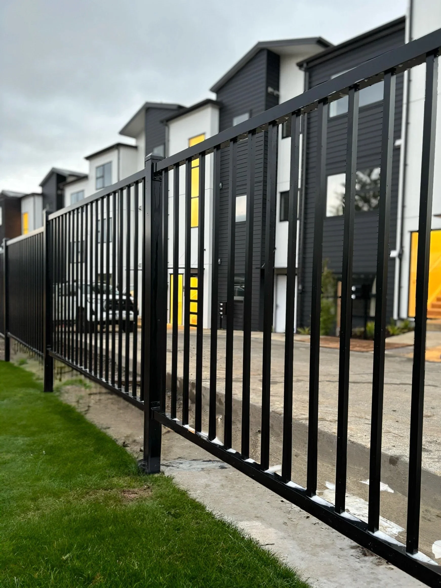 Black metal fence in front of modern multi-story white and dark gray residential buildings with yellow accent panels, over a concrete sidewalk and a patch of green grass, under a cloudy sky.